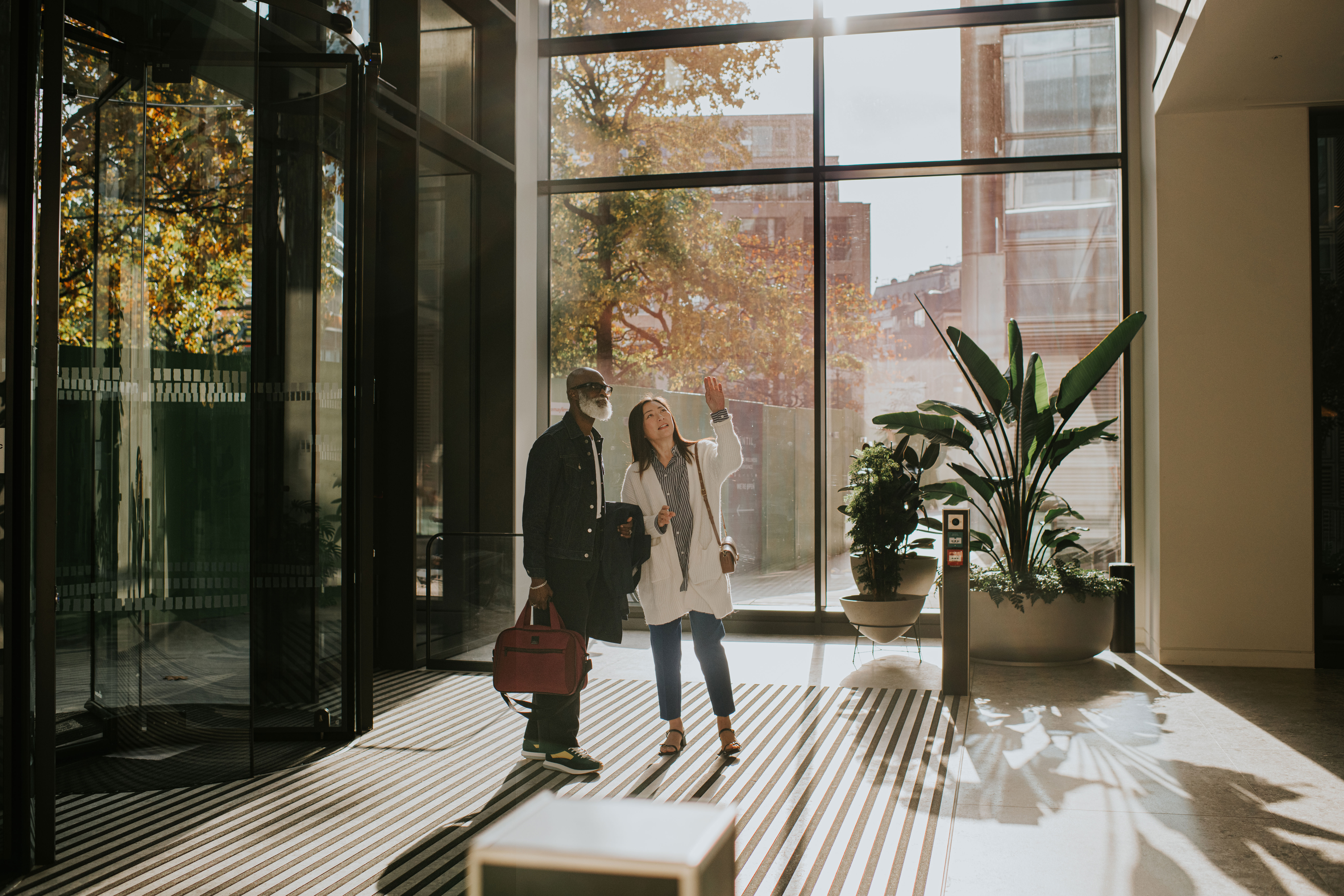 Two people stand in a sunlit, modern building lobby with large windows, one carrying a red bag while the other gestures upward, surrounded by greenery and shadows cast on the floor.