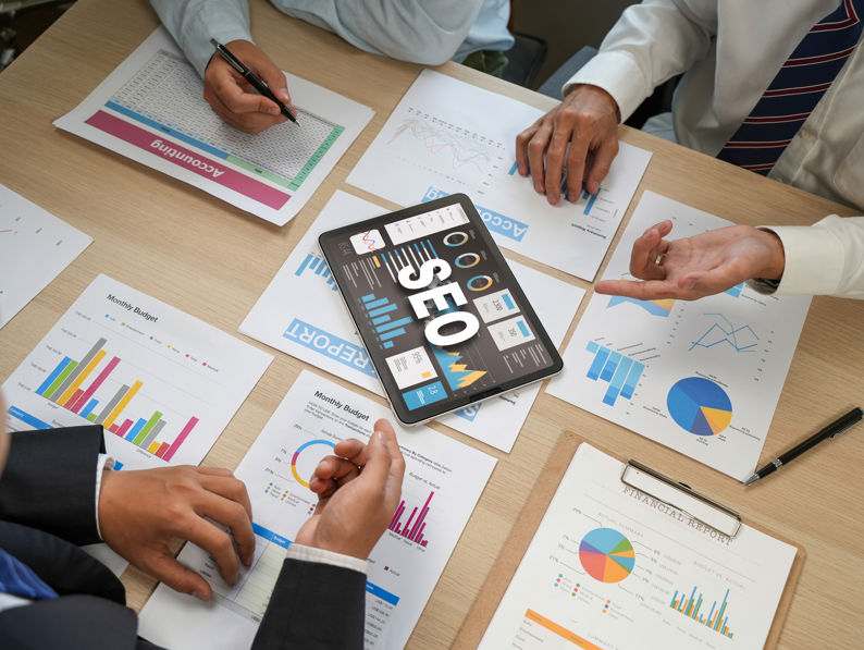 Three people in business attire sit around a table with charts, graphs, and documents. A tablet displaying "SEO" and data visualizations is in the center, as they discuss digital marketing strategies.
