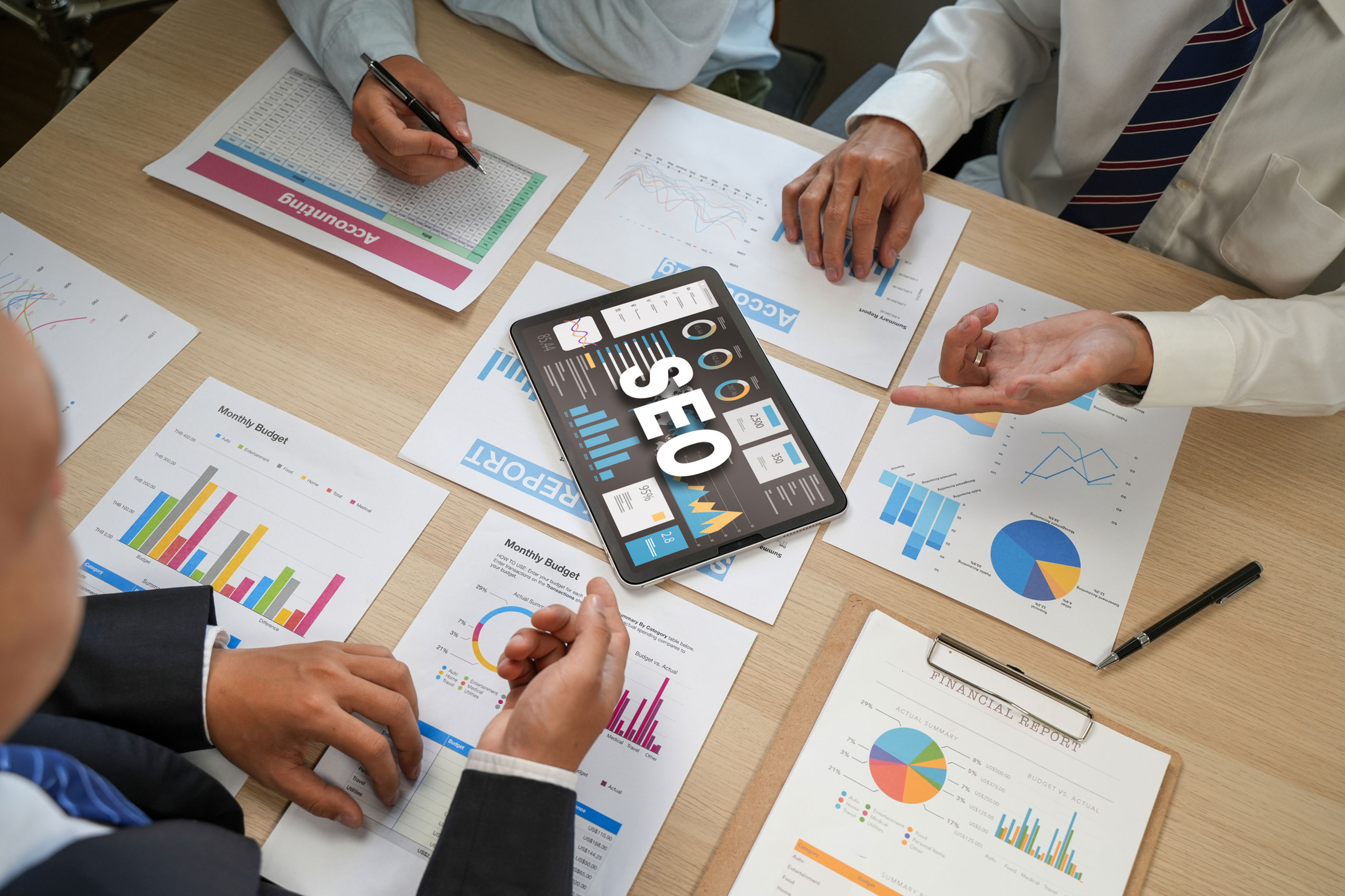 Three people in business attire sit around a table with charts, graphs, and documents. A tablet displaying "SEO" and data visualizations is in the center, as they discuss digital marketing strategies.