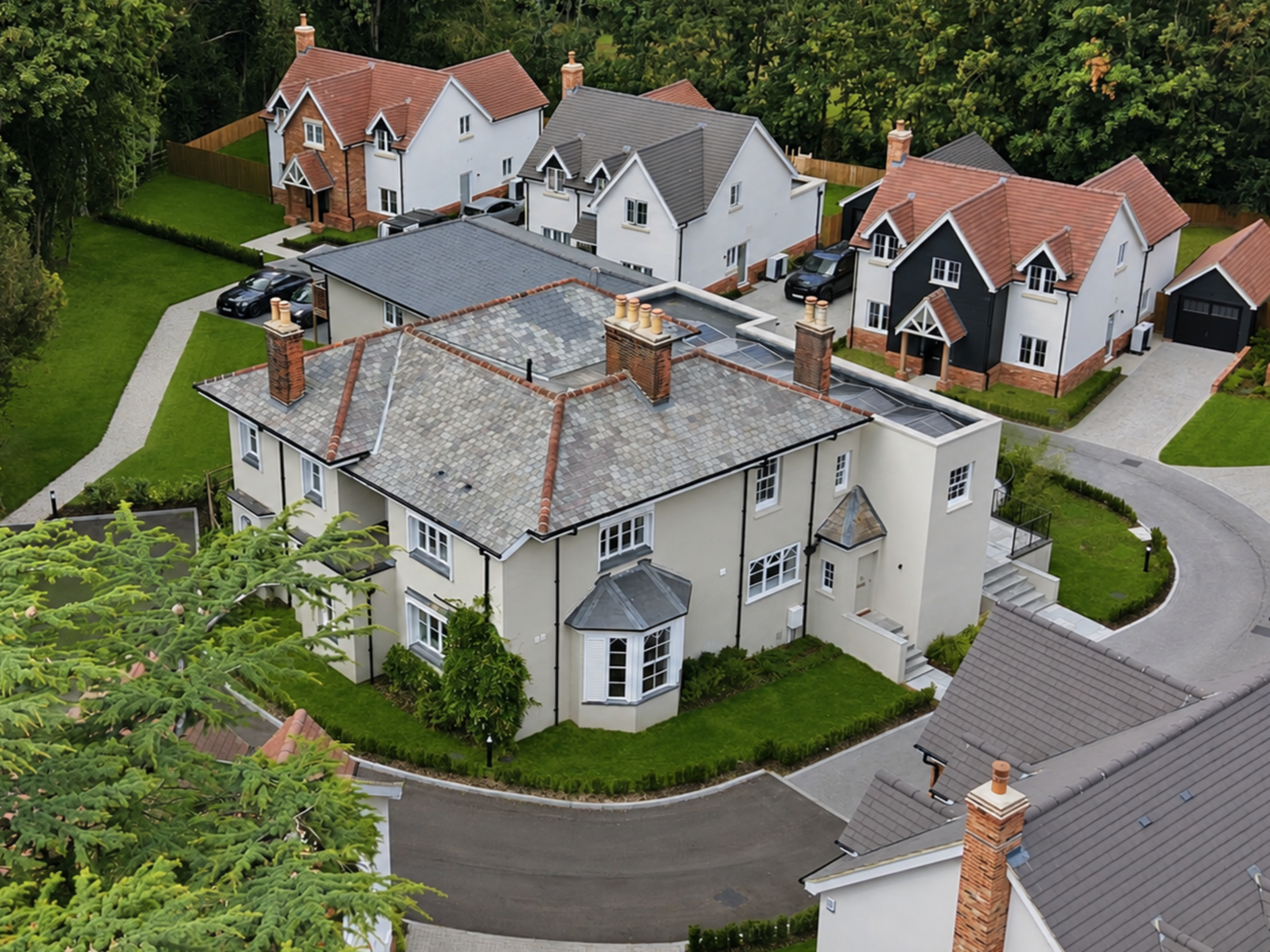 Aerial view of a suburban neighborhood with modern, detached houses, green lawns, driveways, and a curved street surrounded by trees.