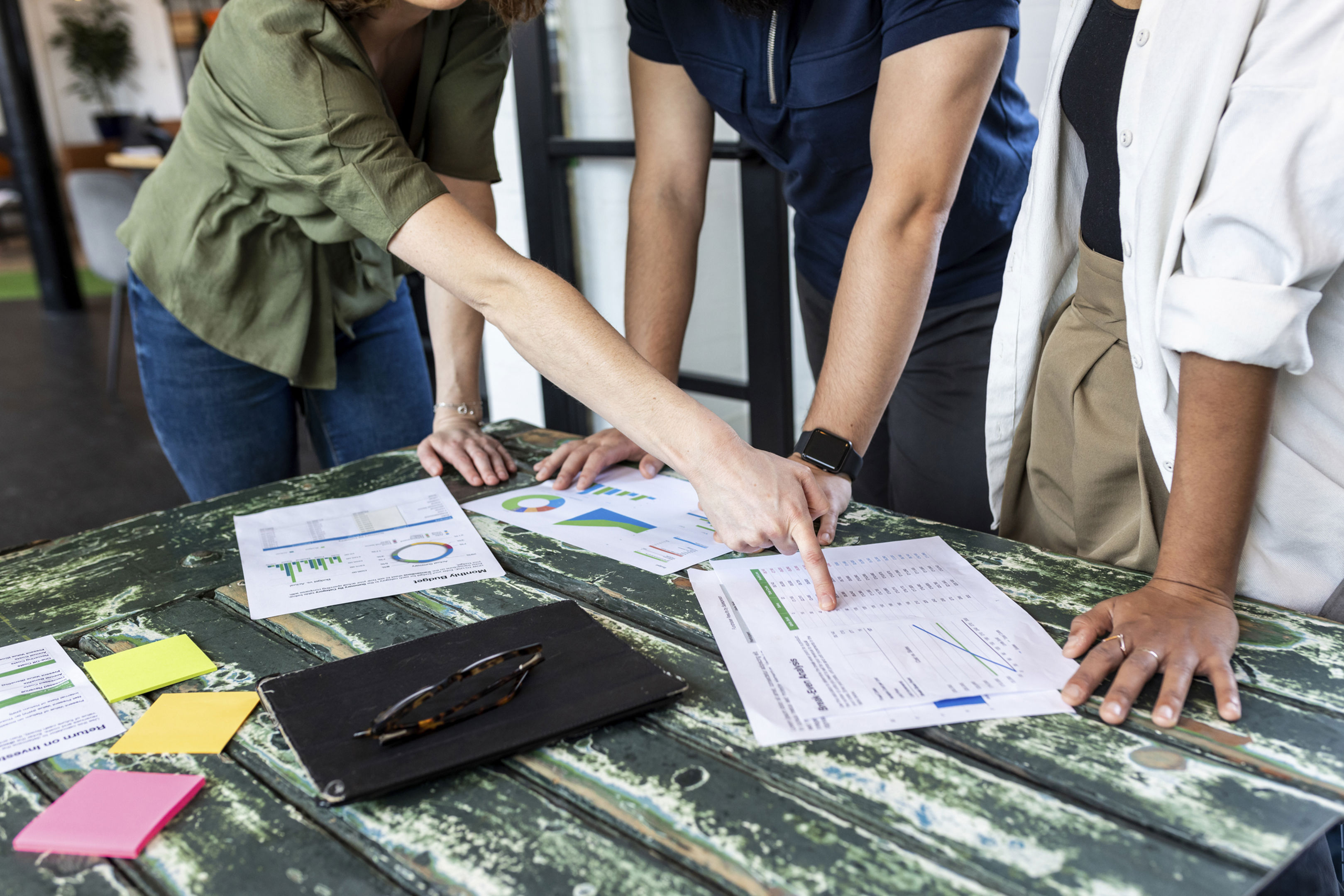 Three people stand around a rustic wooden table, reviewing printed charts and graphs. They are pointing at the documents, which are spread out alongside colorful sticky notes, a notebook, and glasses.