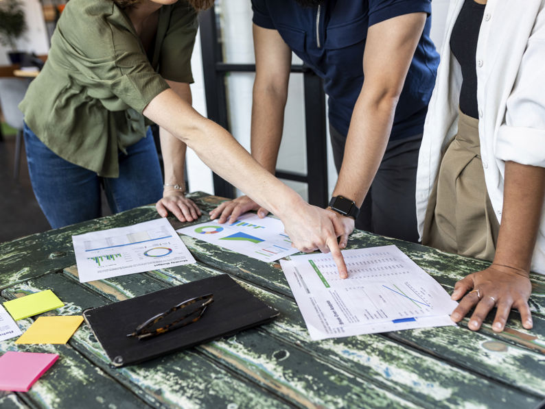 Three people stand around a rustic wooden table, reviewing printed charts and graphs. They are pointing at the documents, which are spread out alongside colorful sticky notes, a notebook, and glasses.