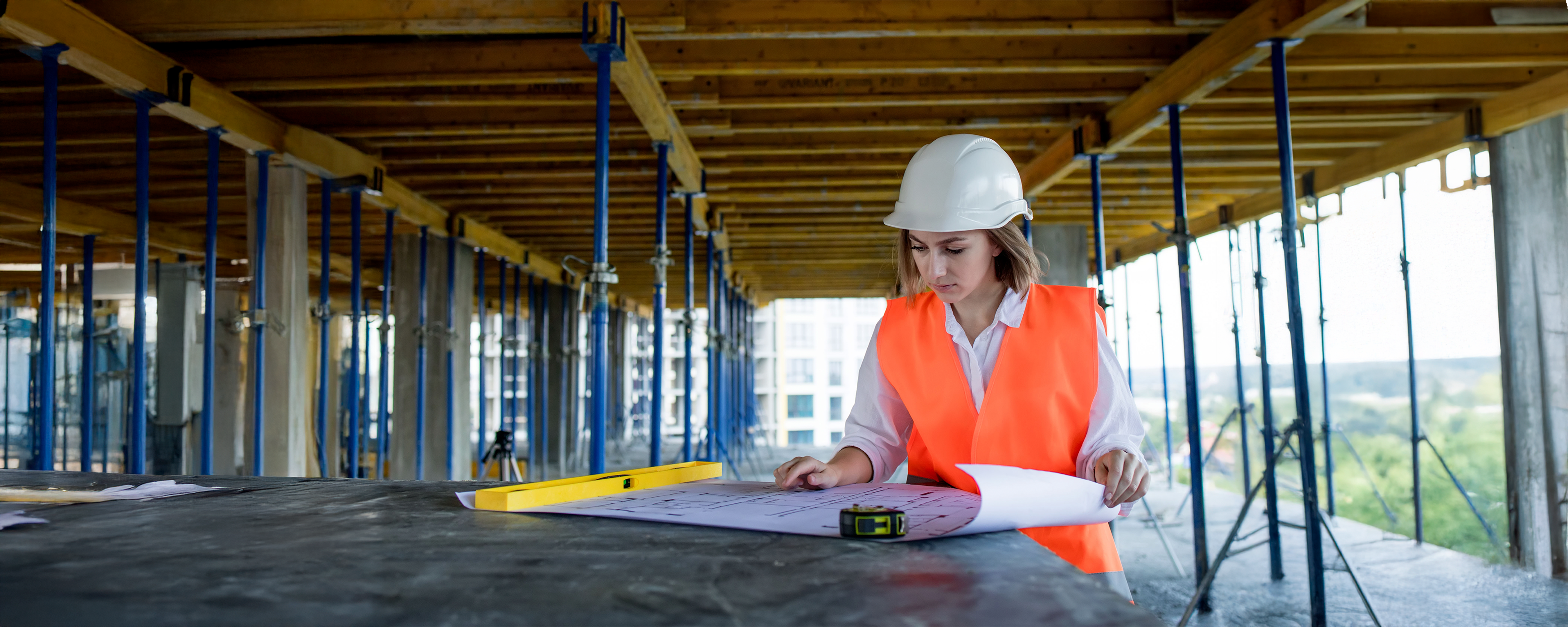 A woman in a white hard hat and orange safety vest examines blueprints on a construction site, surrounded by metal supports and wooden beams.