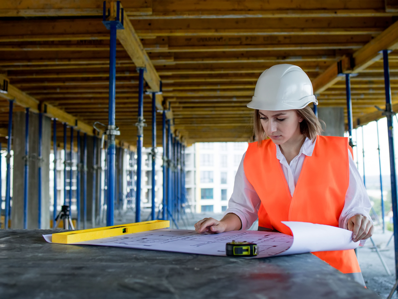 A woman in a white hard hat and orange safety vest examines blueprints on a construction site, surrounded by metal supports and wooden beams.
