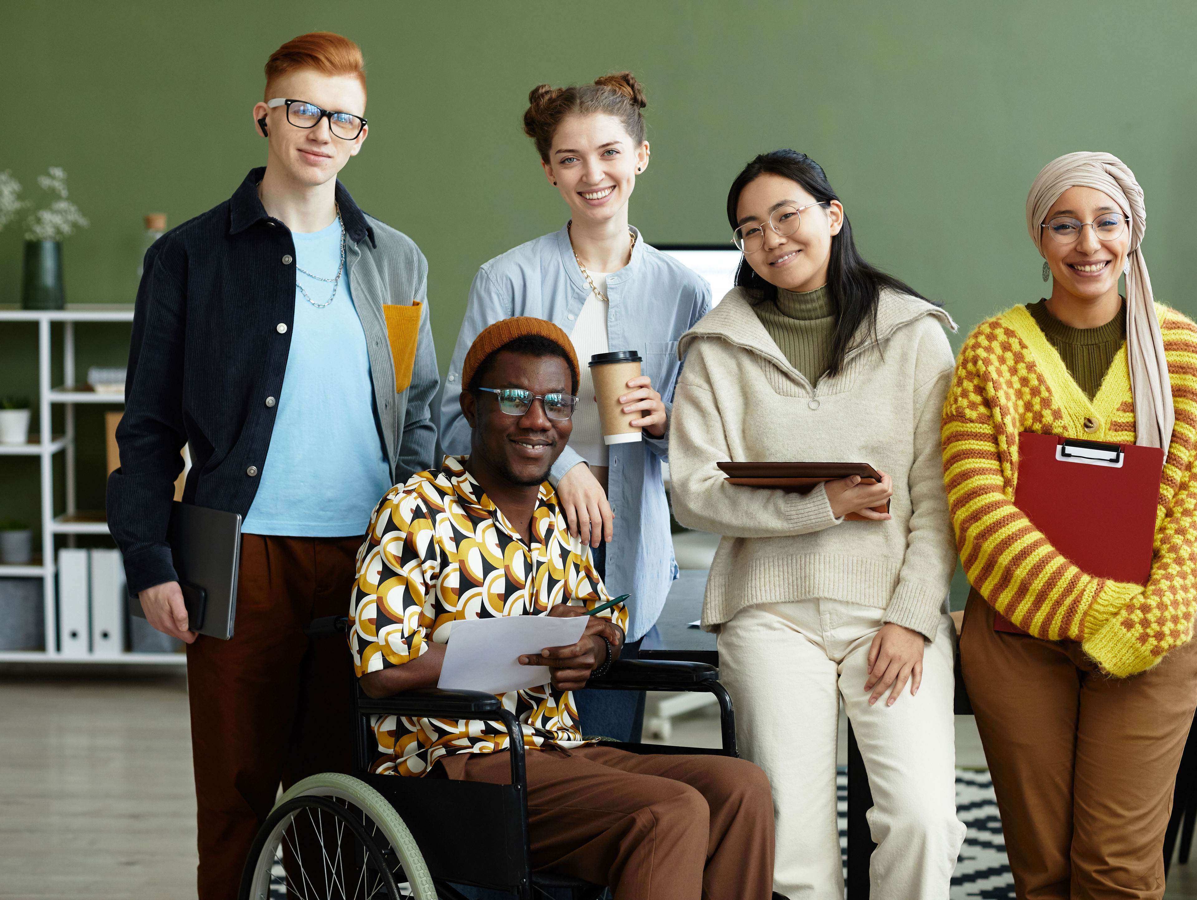 A diverse group of five people smiling indoors. One person is in a wheelchair holding papers, and another holds a coffee cup. They are in a modern office setting with a green wall and bookshelves in the background.