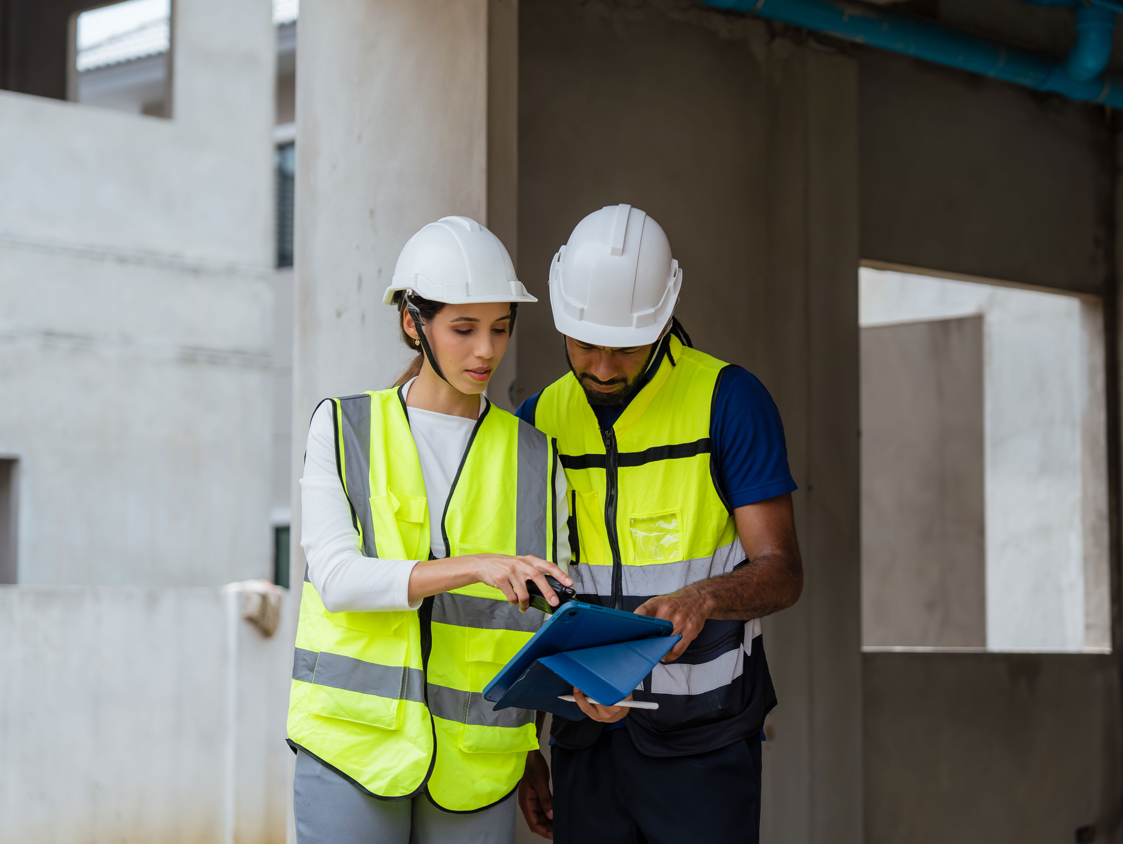 Two construction workers wearing safety vests and helmets review plans on a tablet at a building site, standing near concrete pillars and unfinished walls.