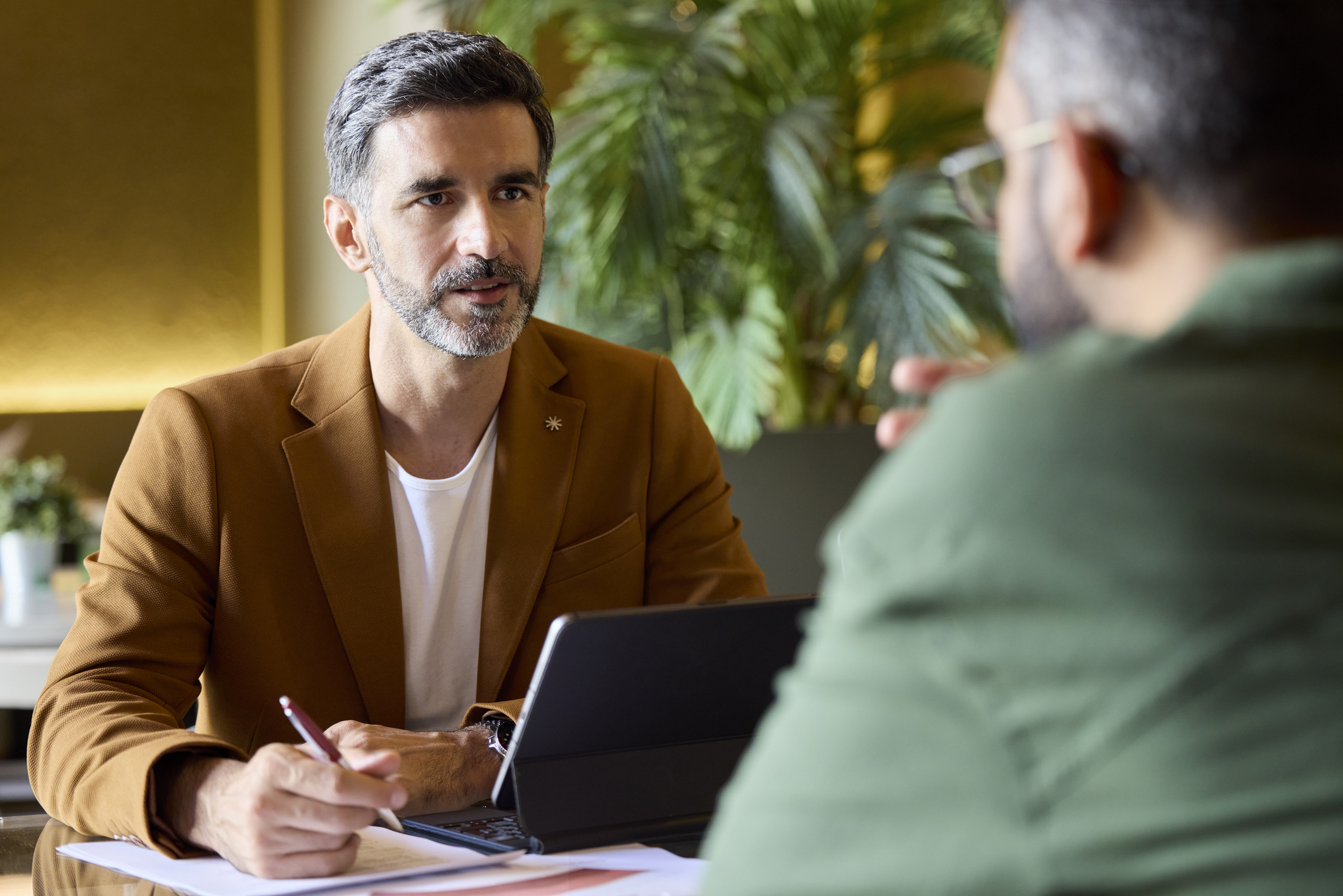 Two men have a serious discussion at a table with a laptop, notepad, and pen. One man in a brown jacket faces the camera, while the other, in green, sits with his back to the viewer. Indoor setting with plants visible.