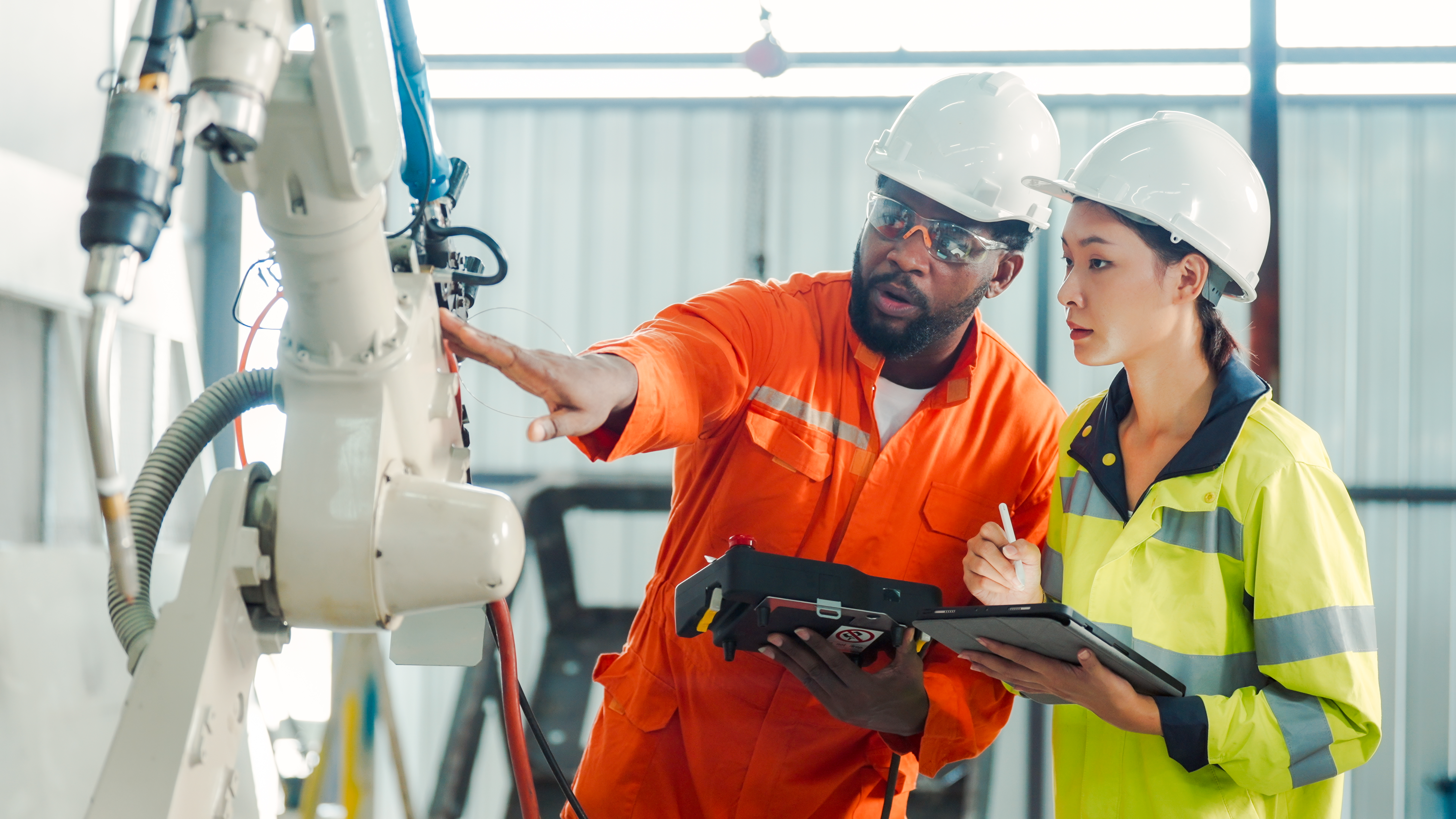 Two engineers in safety helmets and high-visibility clothing examine and discuss a robotic arm in an industrial setting, holding a tablet and clipboard for notes.