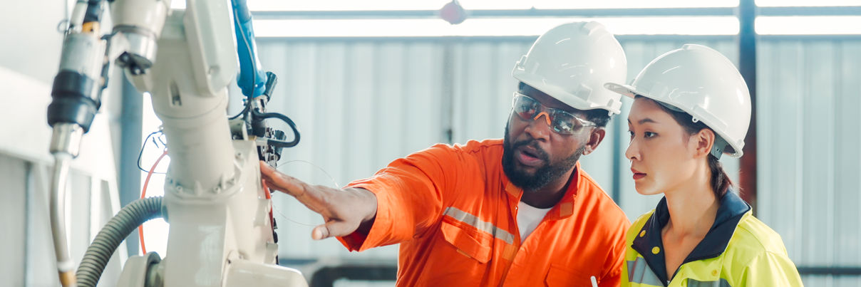 Two engineers in safety helmets and high-visibility clothing examine and discuss a robotic arm in an industrial setting, holding a tablet and clipboard for notes.