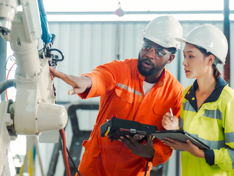 Two engineers in safety helmets and high-visibility clothing examine and discuss a robotic arm in an industrial setting, holding a tablet and clipboard for notes.