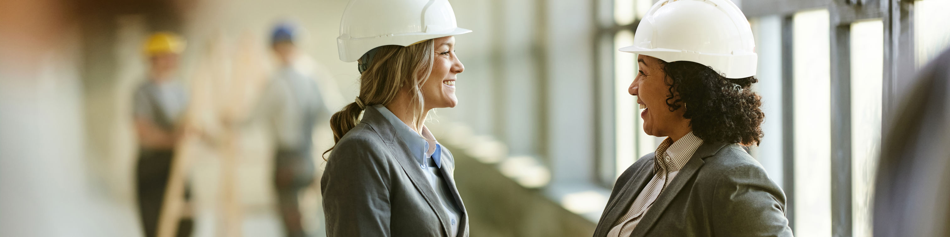 Two women wearing hard hats and business attire shake hands and smile inside a building under construction. Other workers in hard hats are visible in the background near large windows.