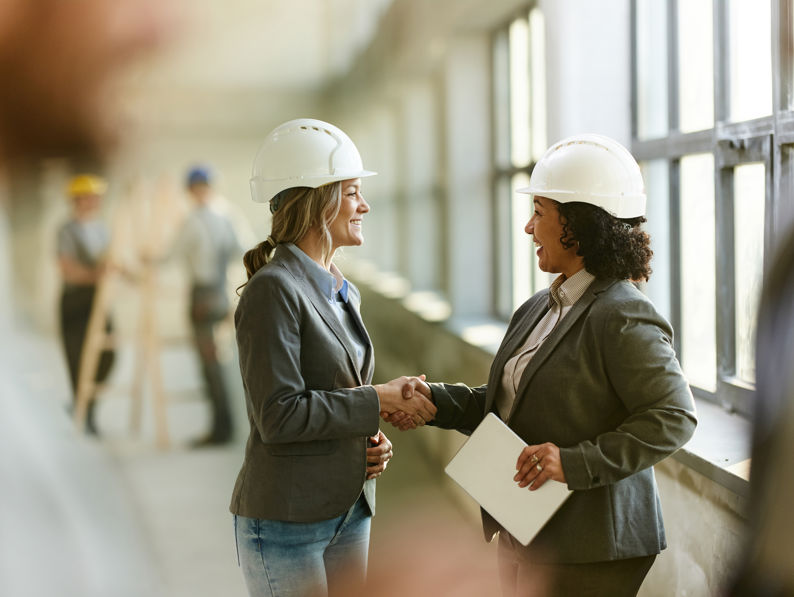 Two women wearing hard hats and business attire shake hands and smile inside a building under construction. Other workers in hard hats are visible in the background near large windows.