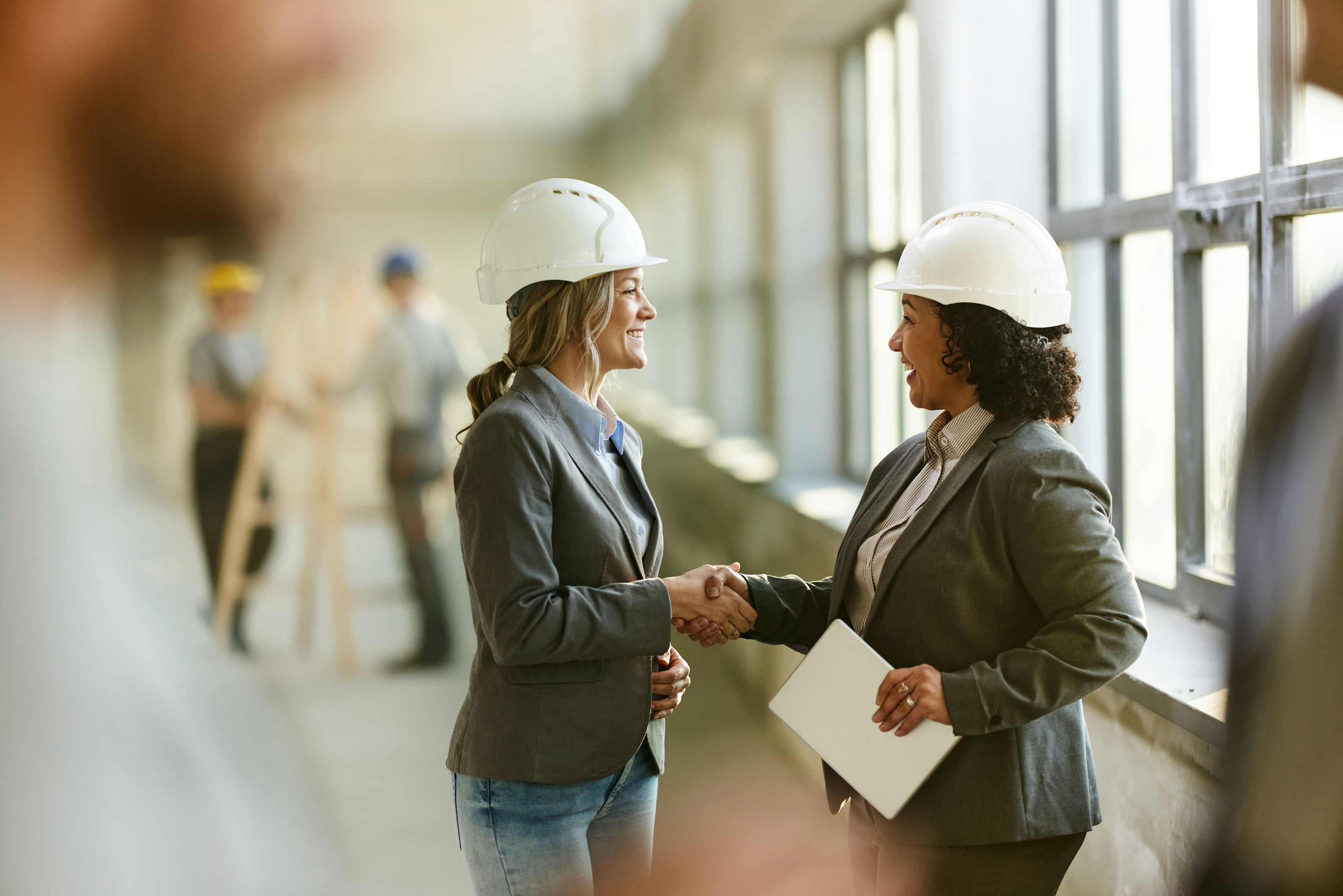 Two women wearing hard hats and business attire shake hands and smile inside a building under construction. Other workers in hard hats are visible in the background near large windows.