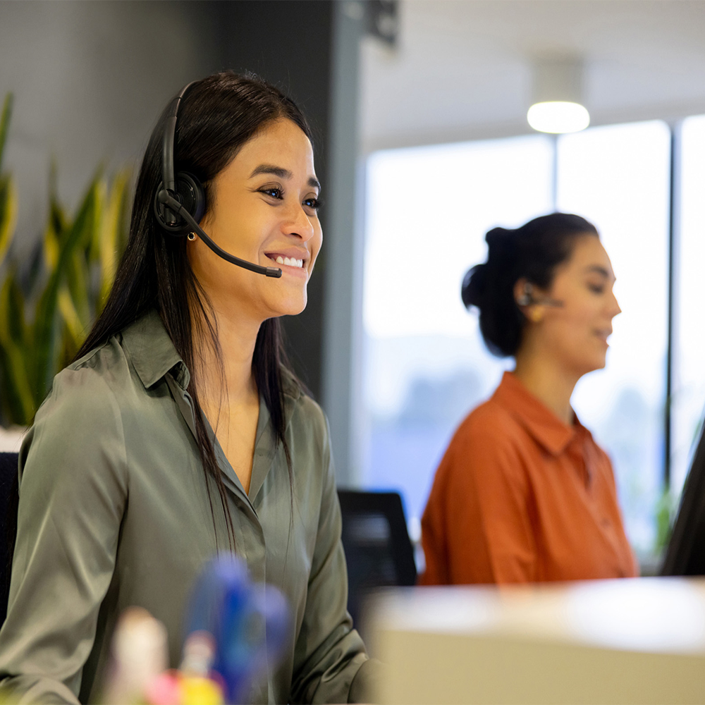 Two women wearing headsets sit at desks in a bright office. The woman in front is smiling and looking at a computer screen, while the woman in the background is focused on her work.
