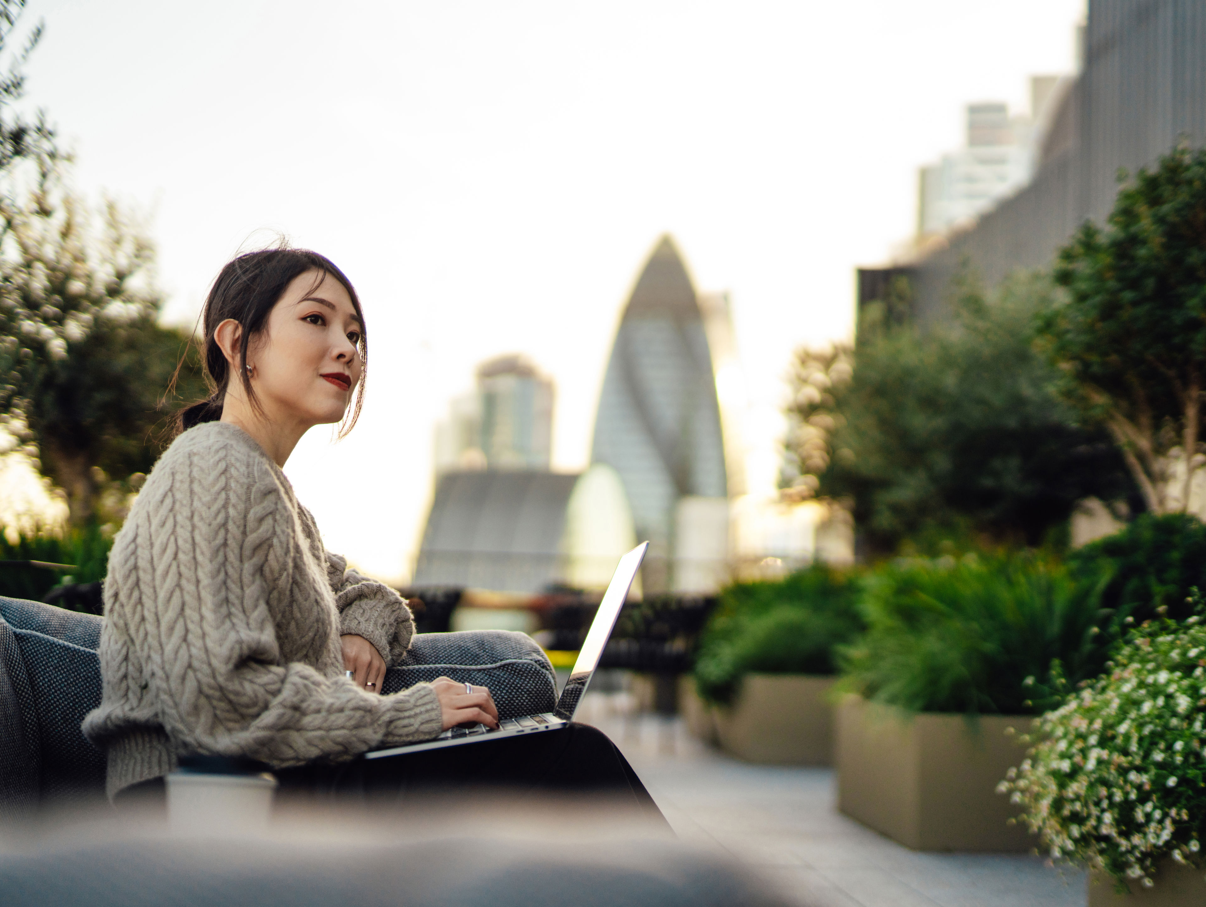 A woman in a beige sweater uses a laptop while sitting outdoors on a city rooftop terrace, with plants around her and tall buildings, including the Gherkin, visible in the background.