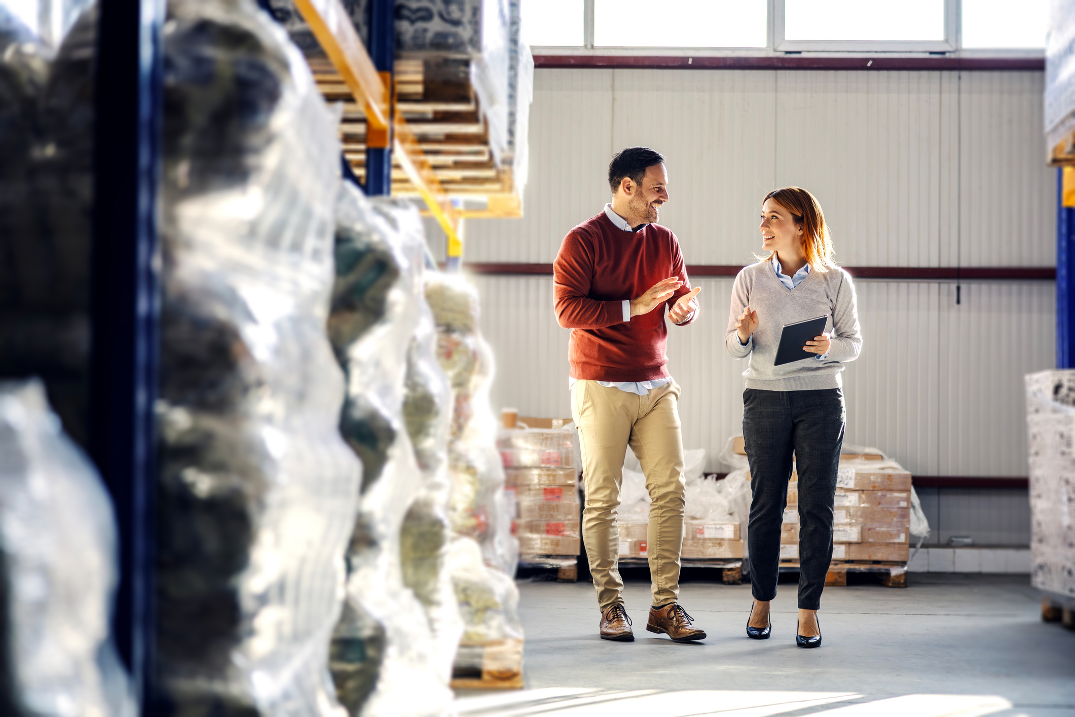 Two people walk and talk in a warehouse filled with stacked pallets and plastic-wrapped goods. One holds a clipboard, and they appear to be having a professional discussion. Sunlight filters through high windows.