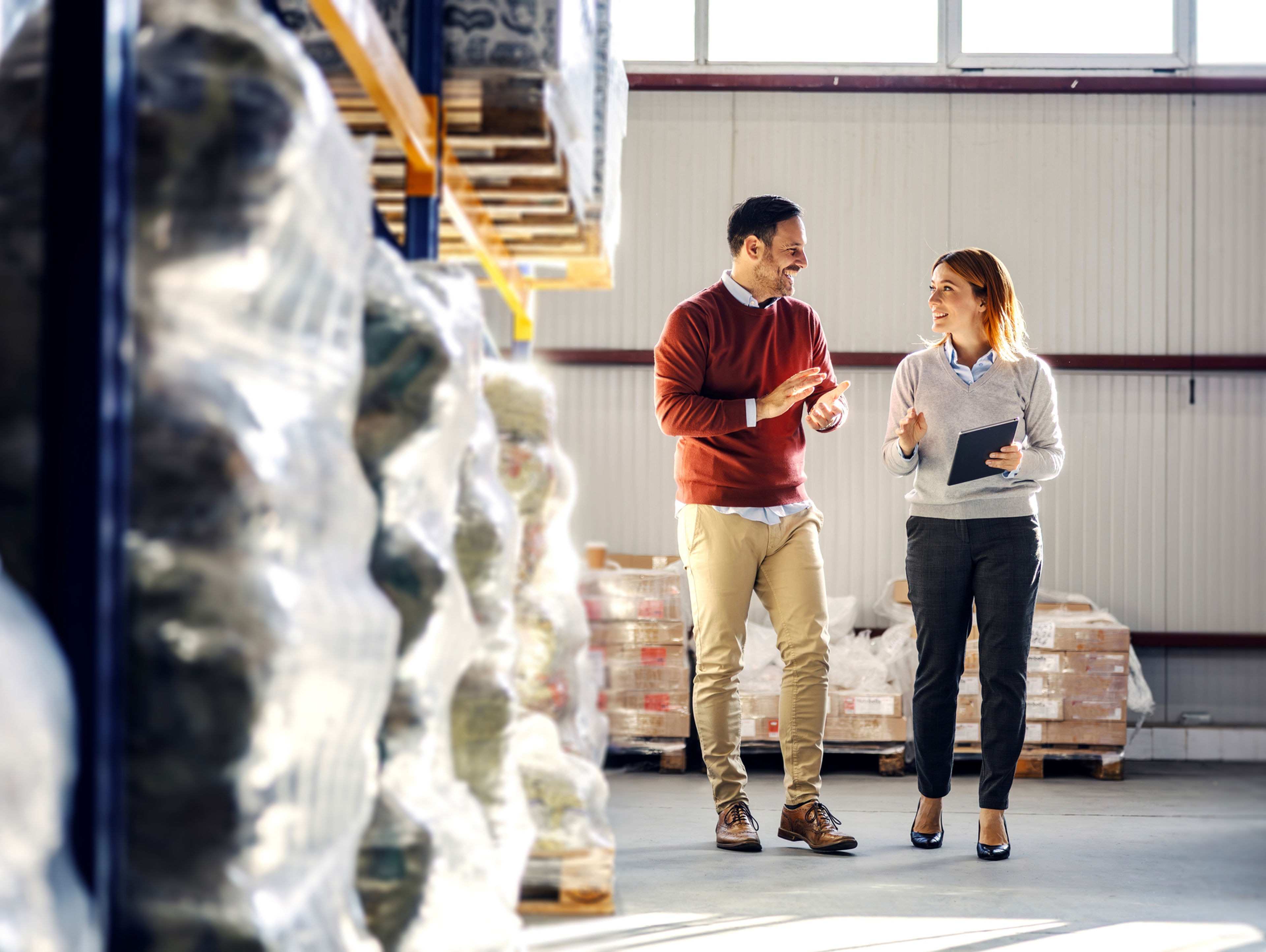 Two people walk and talk in a warehouse filled with stacked pallets and plastic-wrapped goods. One holds a clipboard, and they appear to be having a professional discussion. Sunlight filters through high windows.