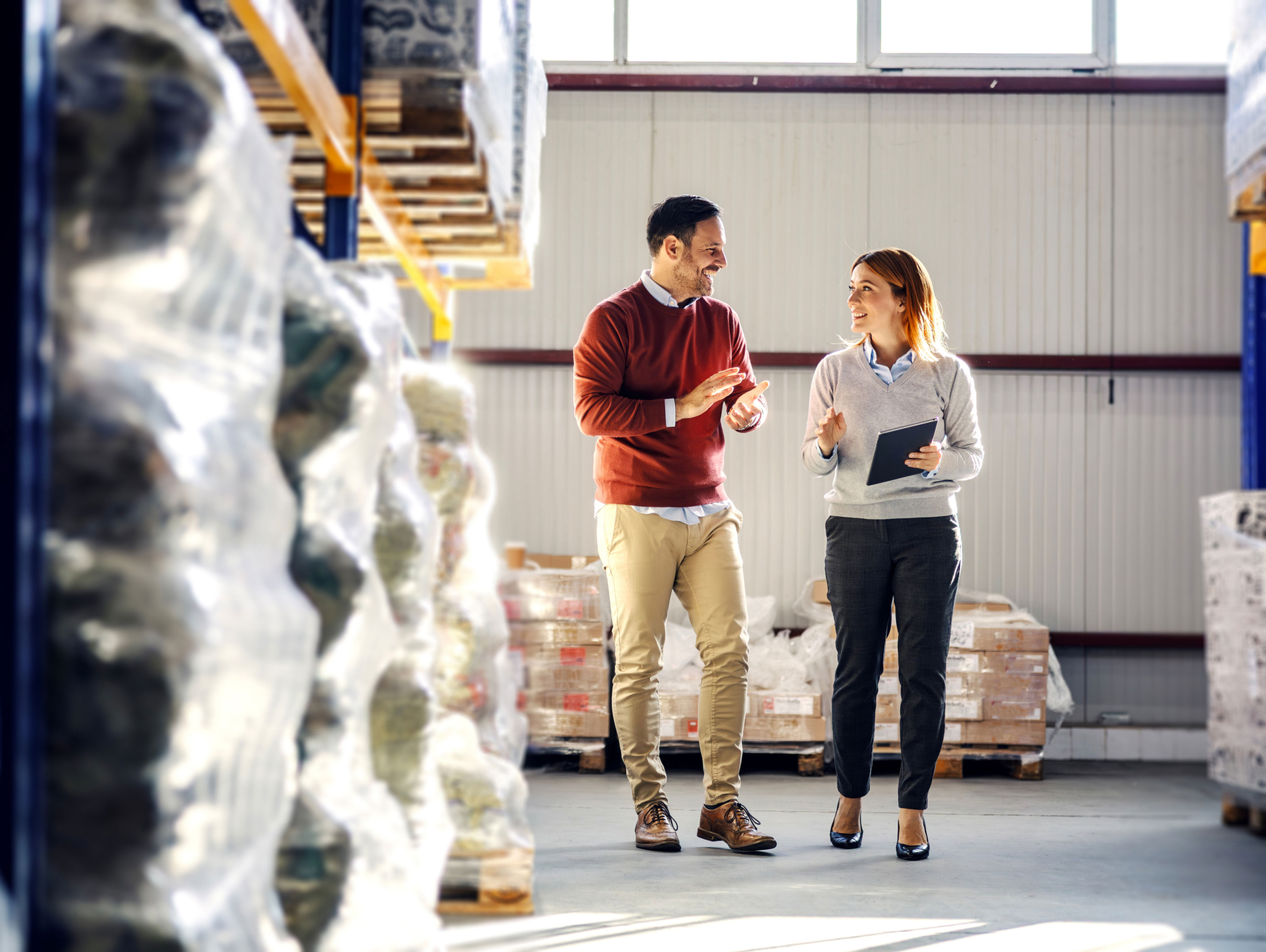 Two people walk and talk in a warehouse filled with stacked pallets and plastic-wrapped goods. One holds a clipboard, and they appear to be having a professional discussion. Sunlight filters through high windows.