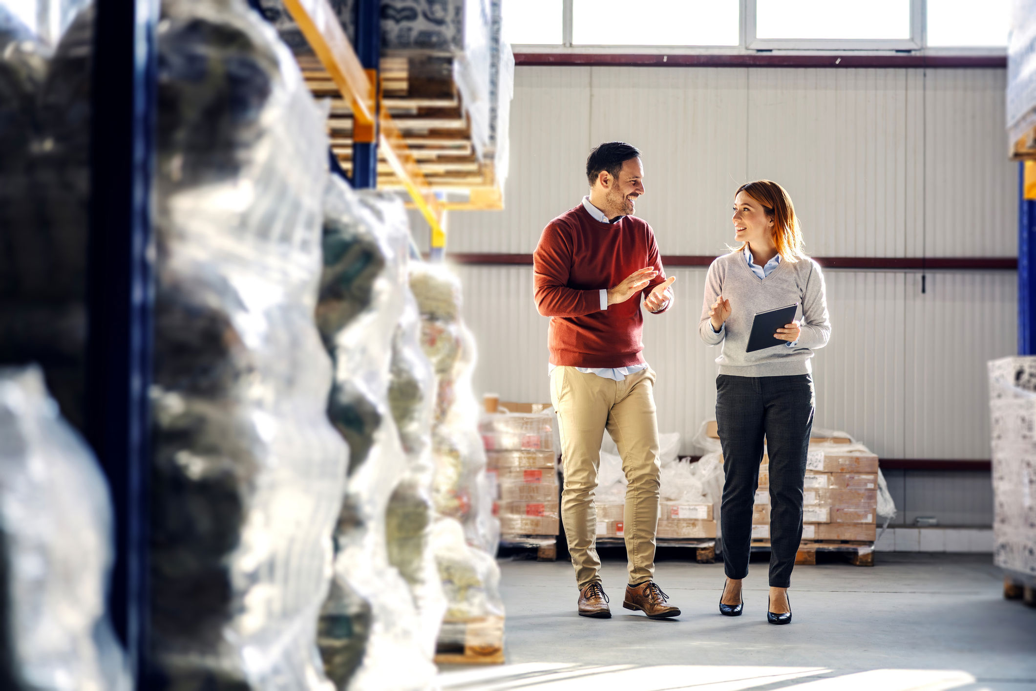 Two people walk and talk in a warehouse filled with stacked pallets and plastic-wrapped goods. One holds a clipboard, and they appear to be having a professional discussion. Sunlight filters through high windows.