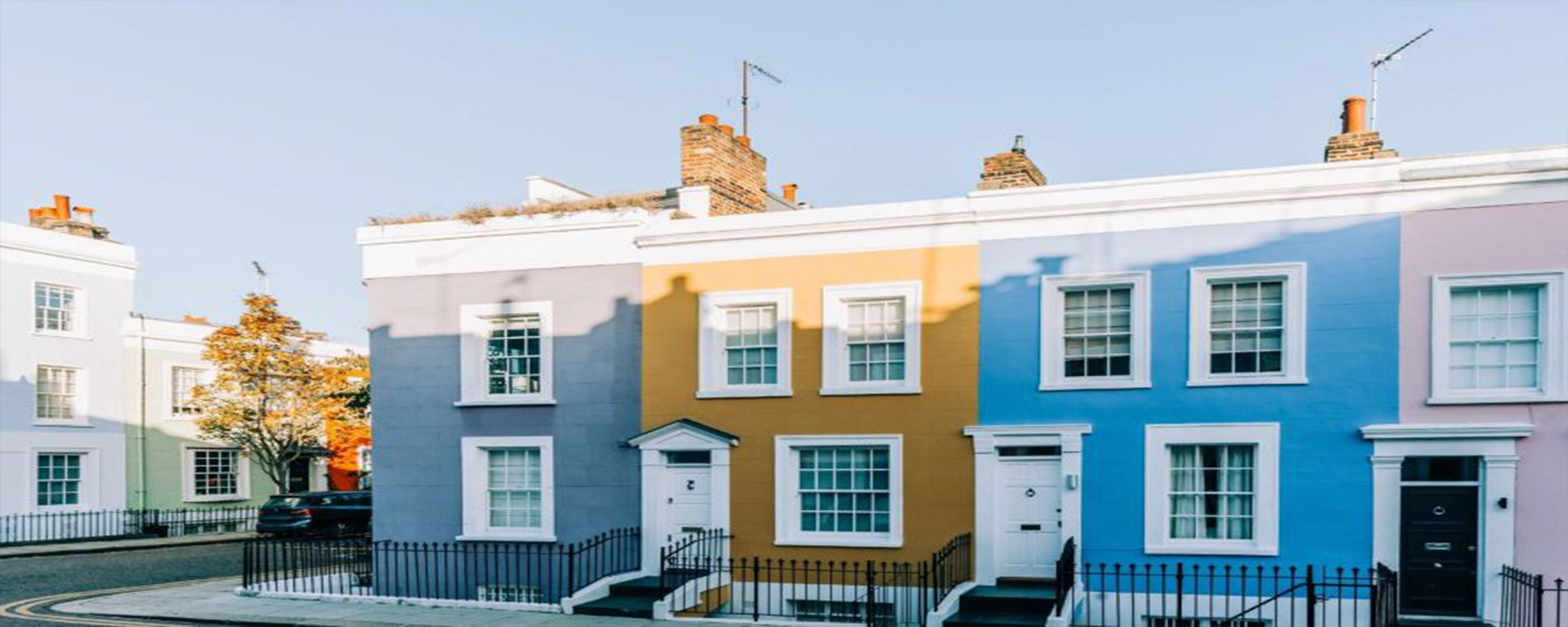 Colorful row houses with white trim, including blue, yellow, and lavender facades, stand side by side on a quiet street under a clear blue sky. A black car is parked on the left near trees with autumn leaves.
