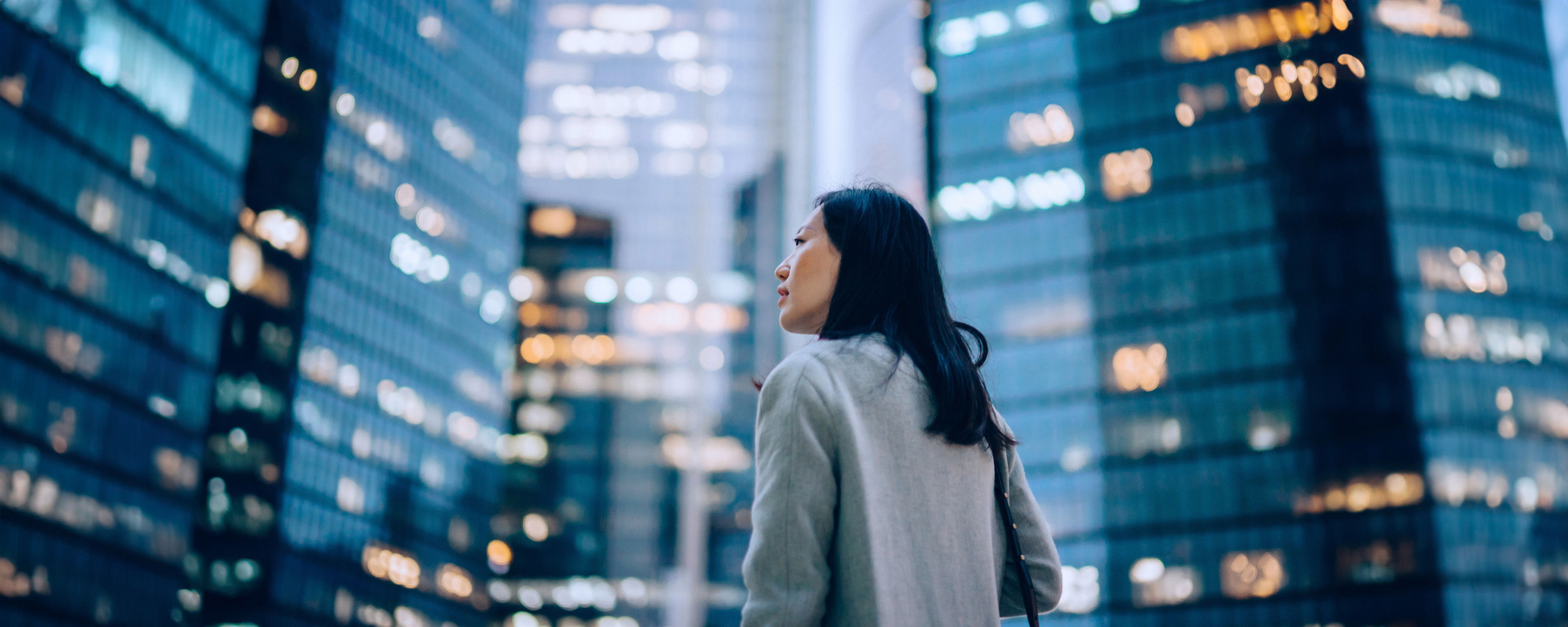 A woman standing in a city street looking up at tall office buildings at dusk.