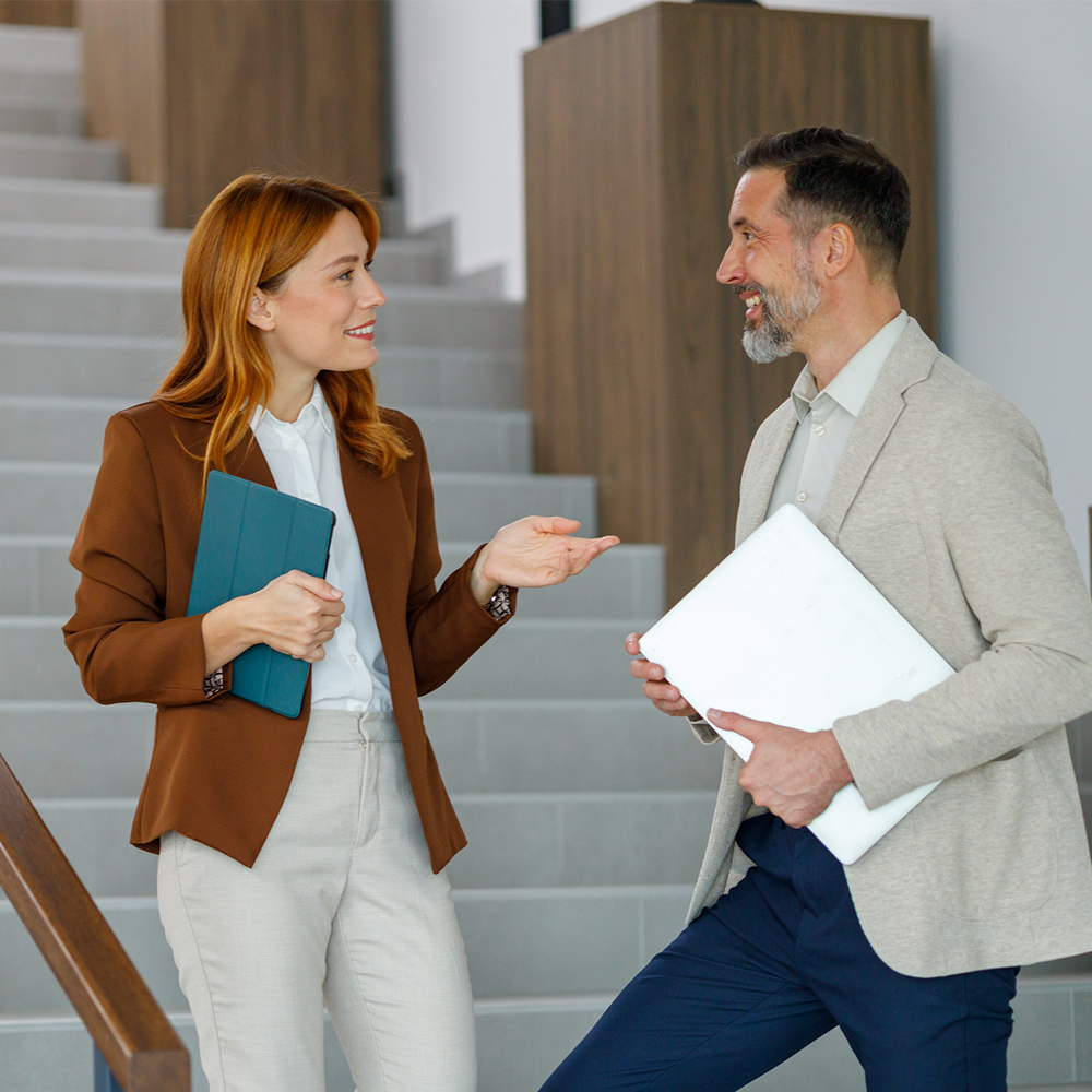 A woman in a brown blazer holding a tablet talks to a man in a beige blazer holding a laptop on a staircase.
