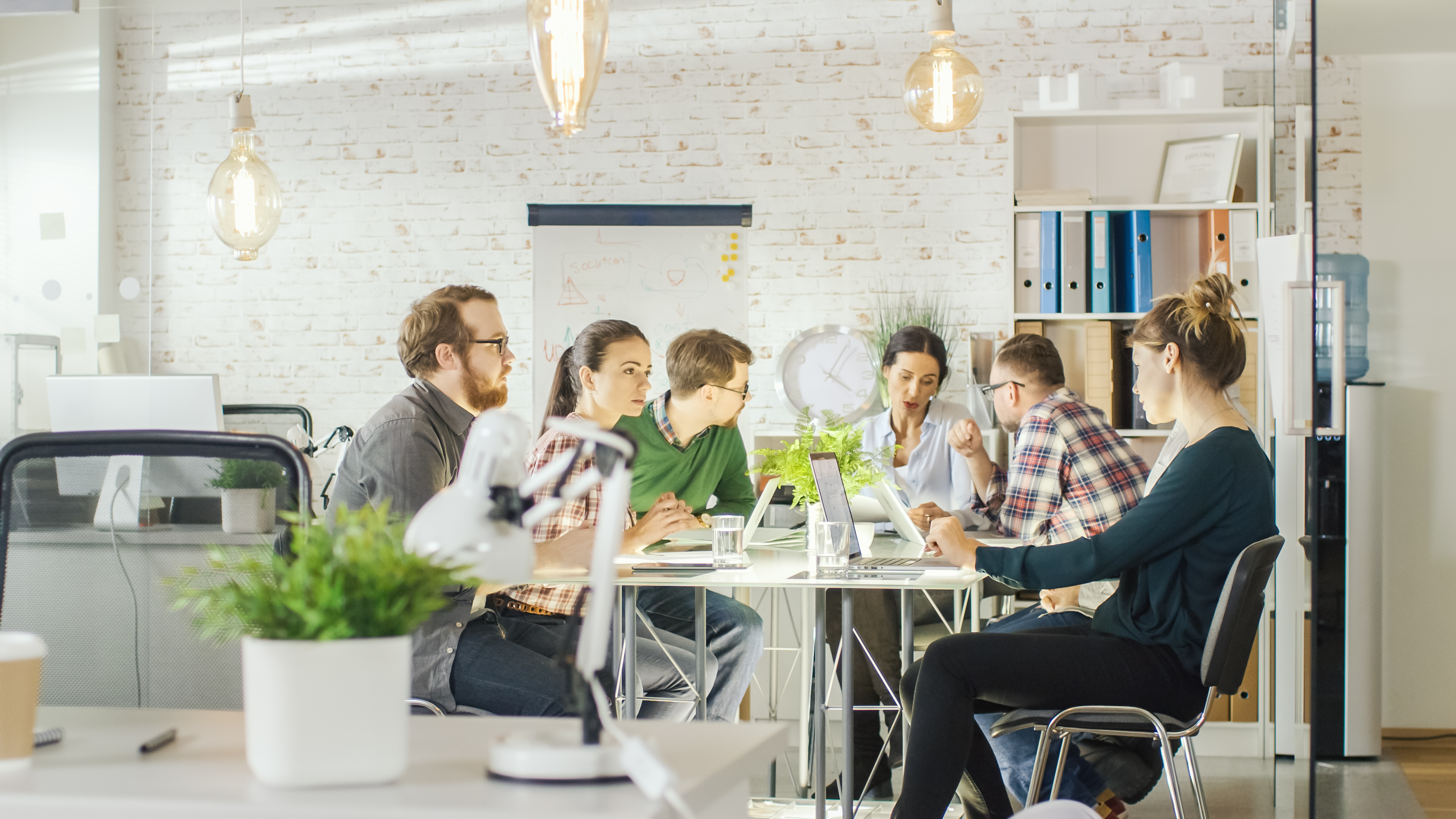 A group of six people sit around a table in a modern, bright office, collaborating and working on laptops, with plants, shelves, and large hanging lights in the background.