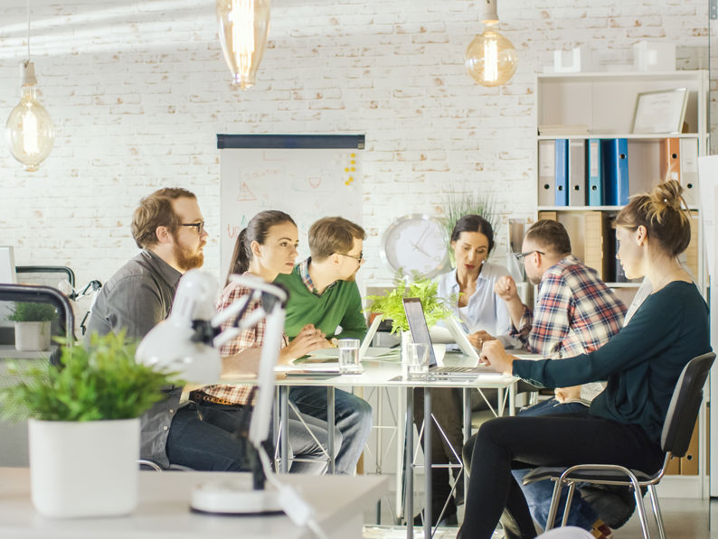 A group of six people sit around a table in a modern, bright office, collaborating and working on laptops, with plants, shelves, and large hanging lights in the background.