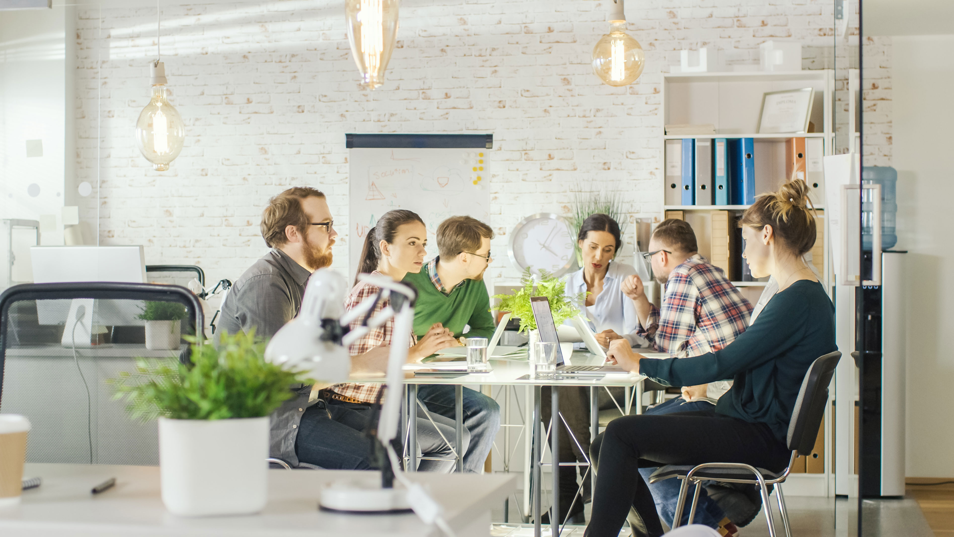 A group of six people sit around a table in a modern, bright office, collaborating and working on laptops, with plants, shelves, and large hanging lights in the background.