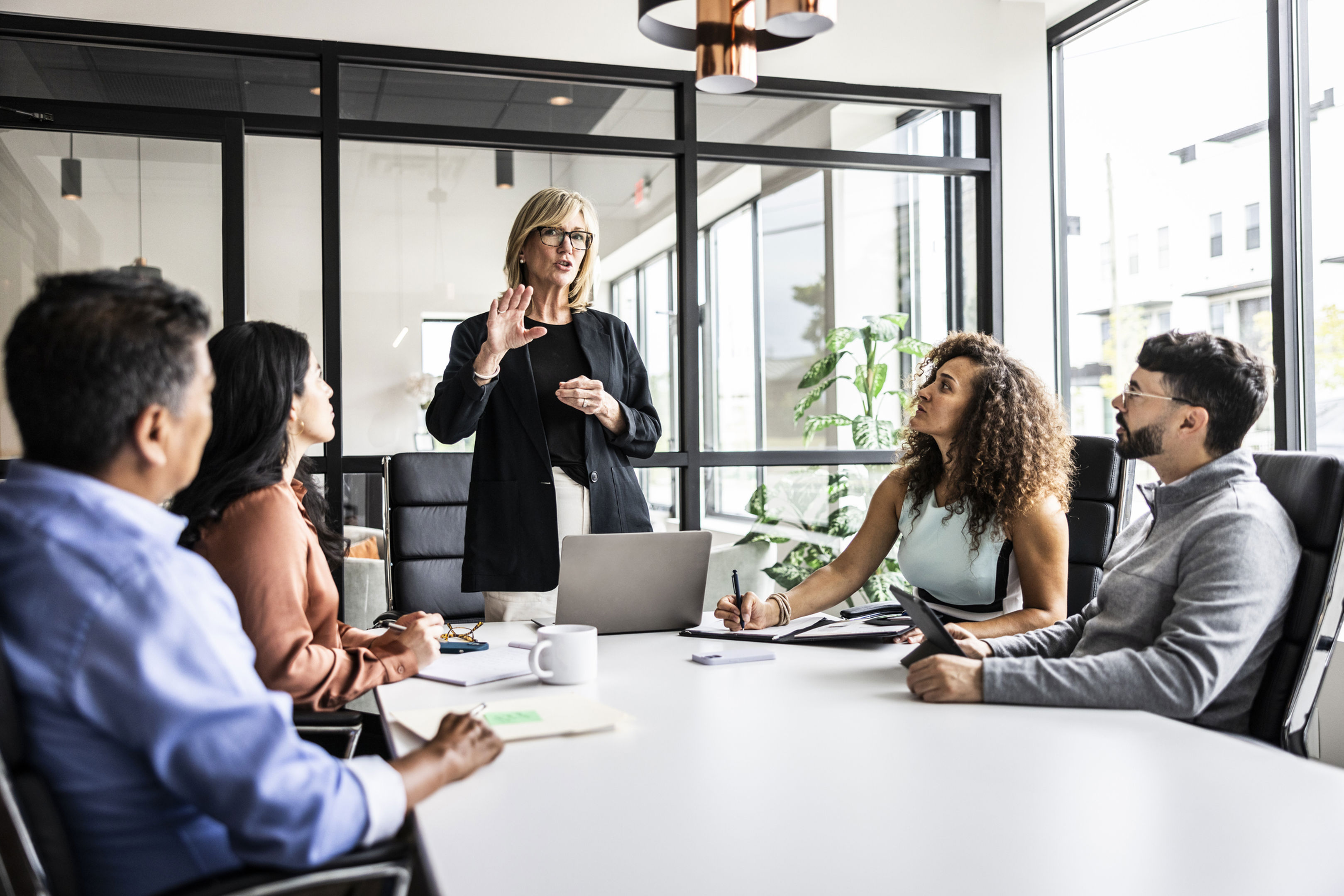 A woman stands addressing a group of four people seated around a conference table in a modern office. One person is taking notes, others listen attentively. A glass wall and potted plant are in the background.