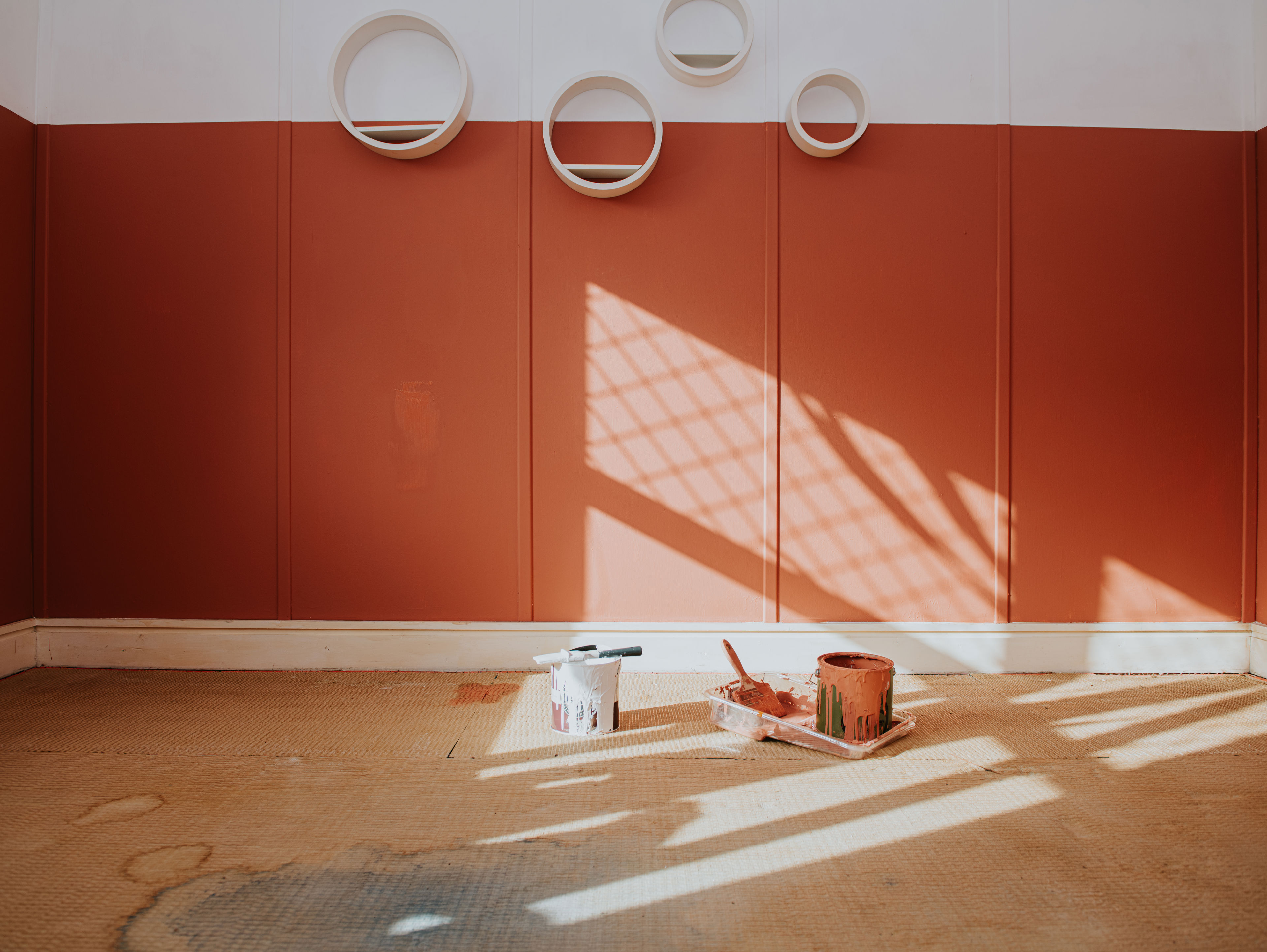 A room with red and white walls, circular shelves on the upper wall, and painting supplies including paint cans, brushes, and a tray on the floor; sunlight casts shadows across the space.