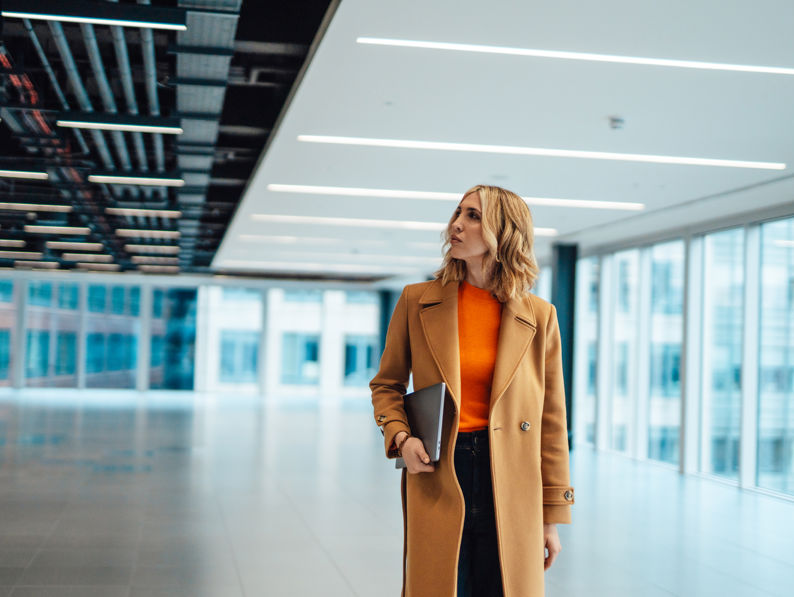 A woman in a brown coat and orange top holds a laptop while walking through a modern, spacious office building with large windows and bright lighting.