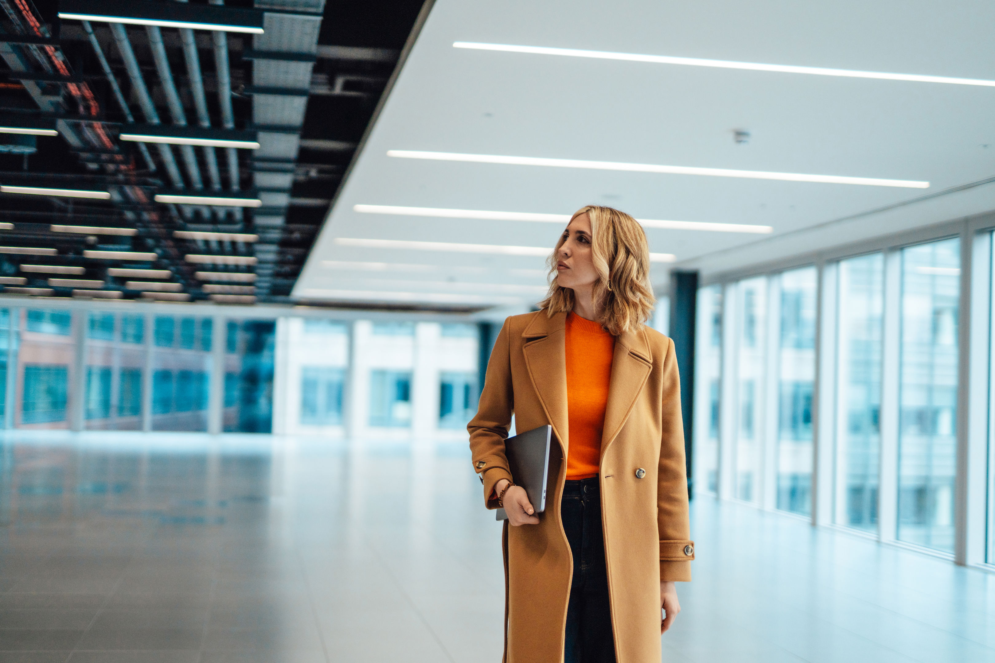 A woman in a brown coat and orange top holds a laptop while walking through a modern, spacious office building with large windows and bright lighting.