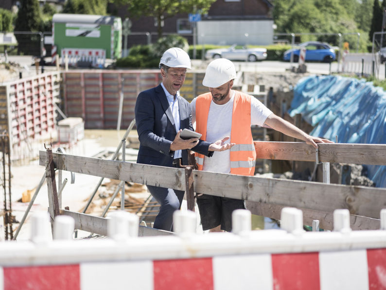 A man in a suit and hard hat shows a tablet to a construction worker wearing a safety vest and helmet at a building site, with equipment and barriers in the background.