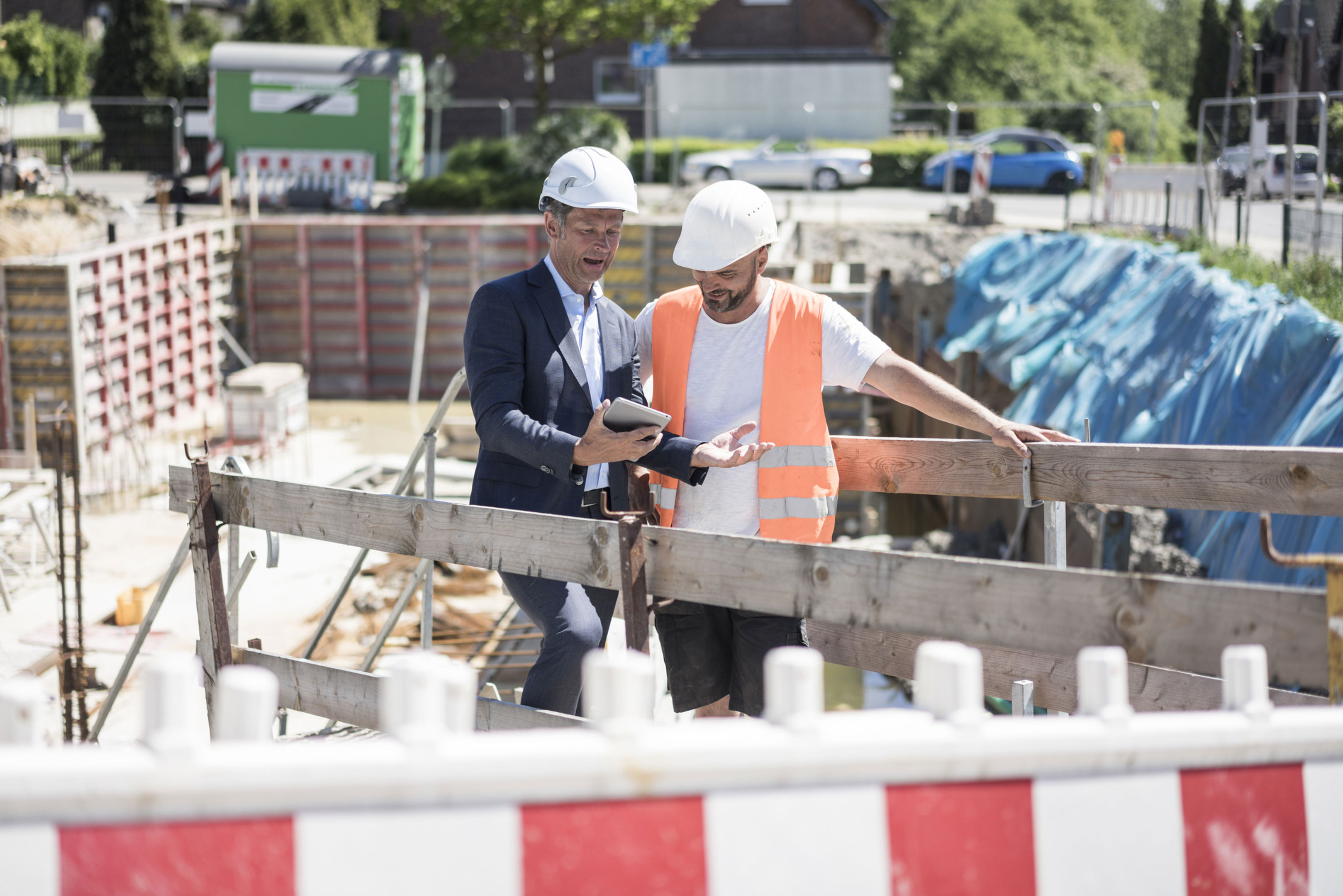 A man in a suit and hard hat shows a tablet to a construction worker wearing a safety vest and helmet at a building site, with equipment and barriers in the background.