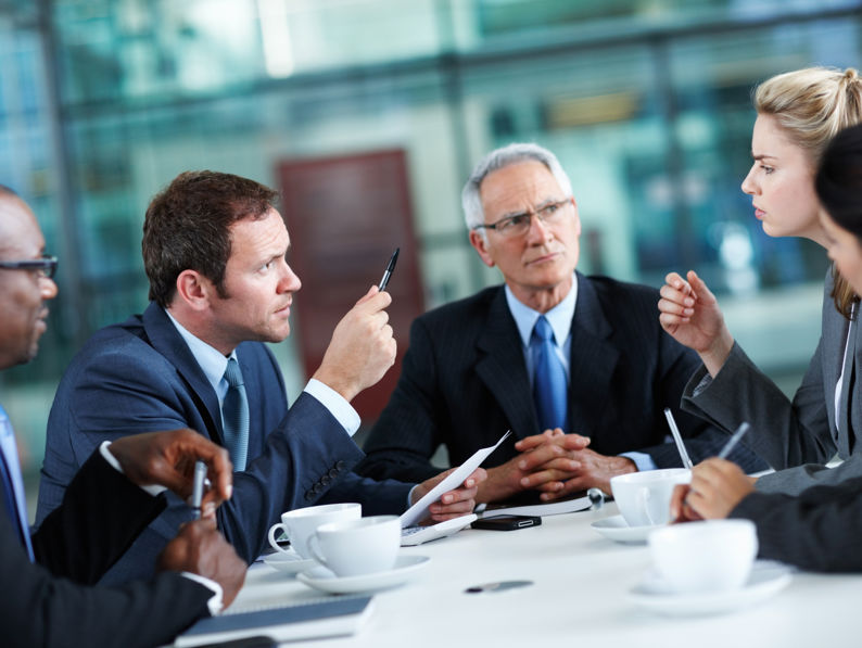 Four business professionals in suits sit around a table having a serious discussion, with one man gesturing with a pen and a woman speaking while others listen attentively. Coffee cups and documents are visible on the table.