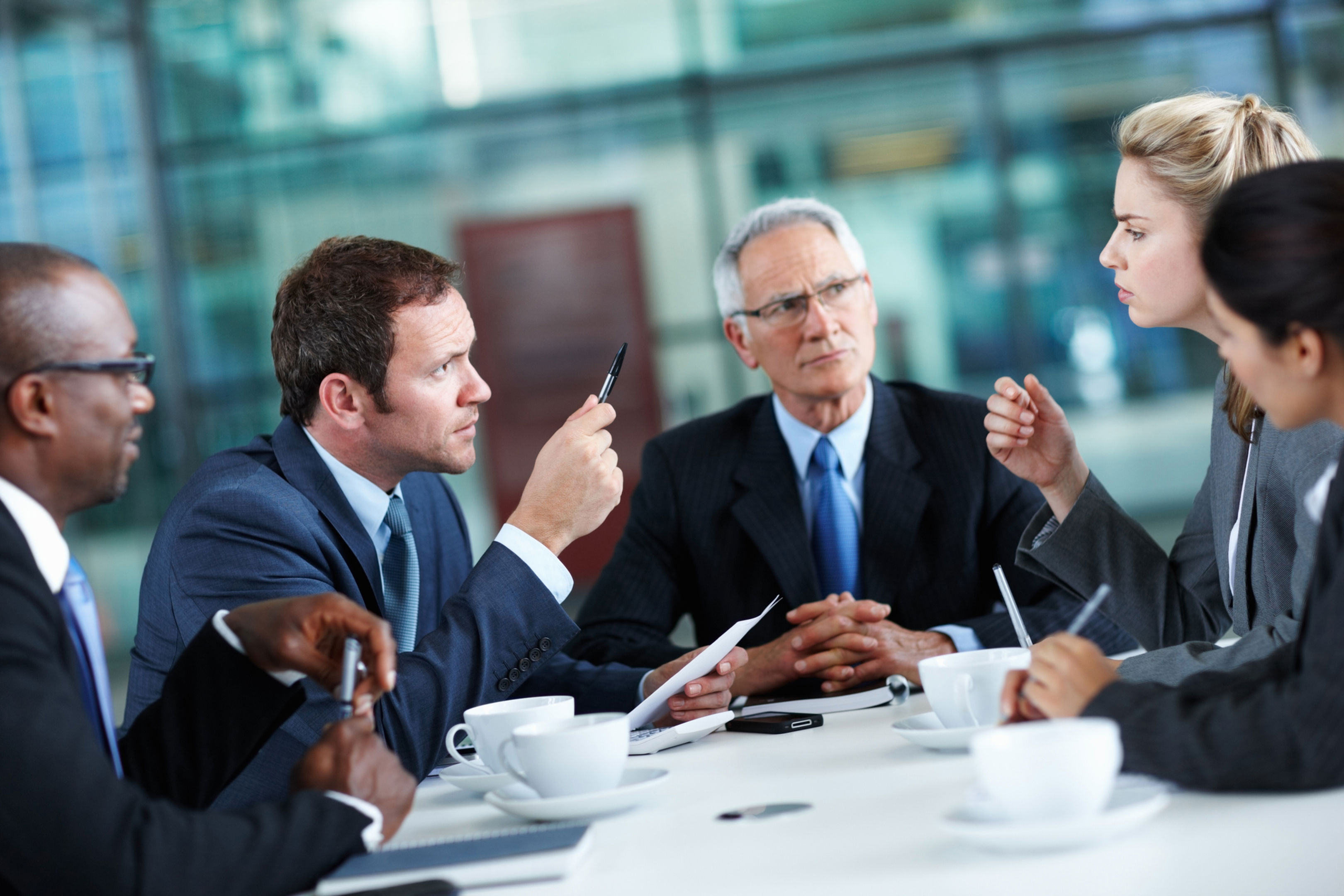 Four business professionals in suits sit around a table having a serious discussion, with one man gesturing with a pen and a woman speaking while others listen attentively. Coffee cups and documents are visible on the table.