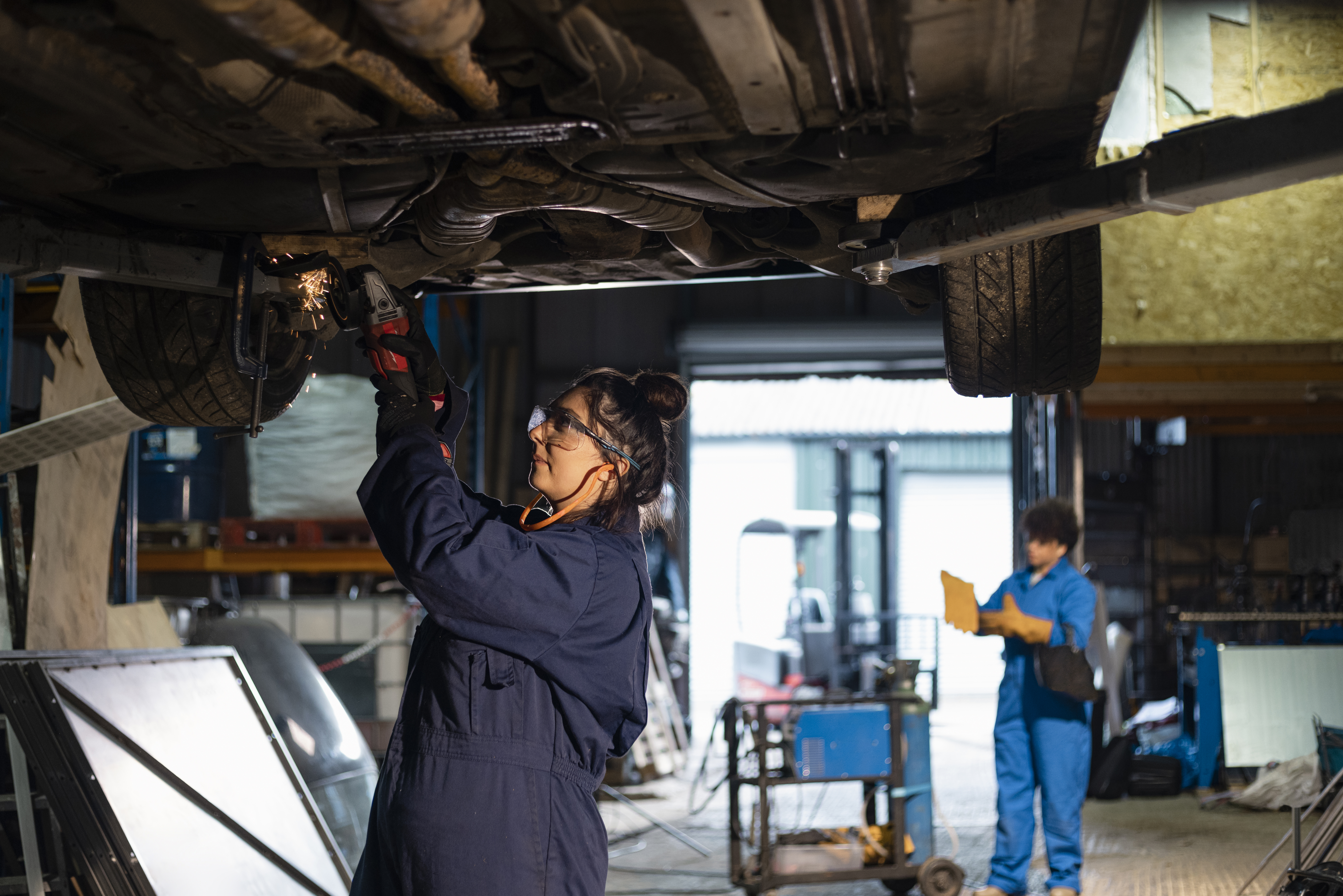 A mechanic in coveralls and safety glasses works under a lifted car with sparks flying, while another person in blue coveralls holds a clipboard in the background of a garage workshop.