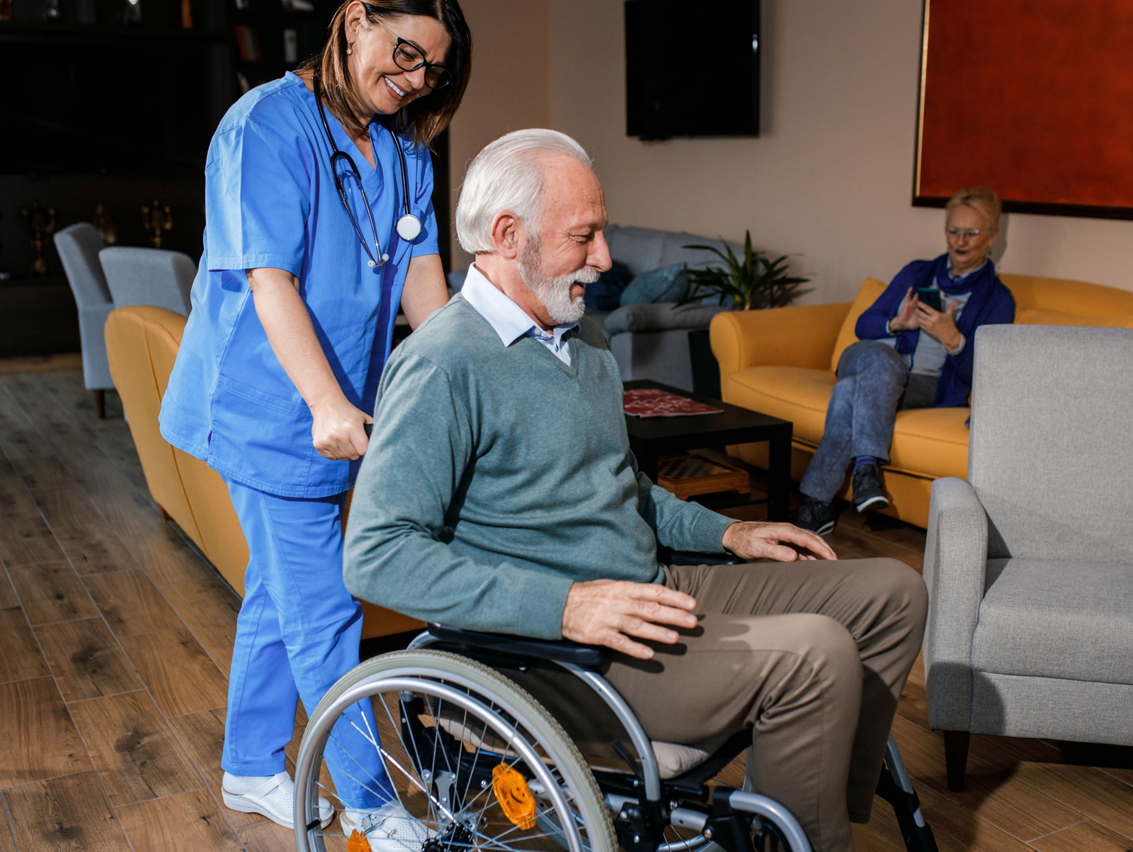 A nurse in blue scrubs smiles while pushing an elderly man in a wheelchair through a living room area; another elderly person sits on a sofa in the background, reading or using a device.