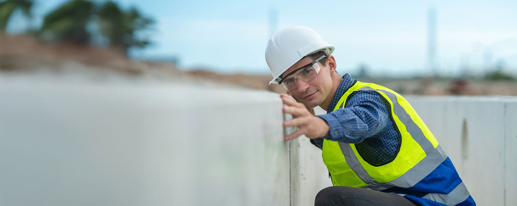 A man in a hard hat with safety glasses on crouched touching a wall