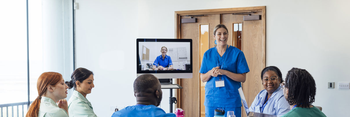 A group of healthcare professionals sit around a table, listening to a colleague in blue scrubs who is standing and presenting. Another person in scrubs appears on a monitor, participating via video call.