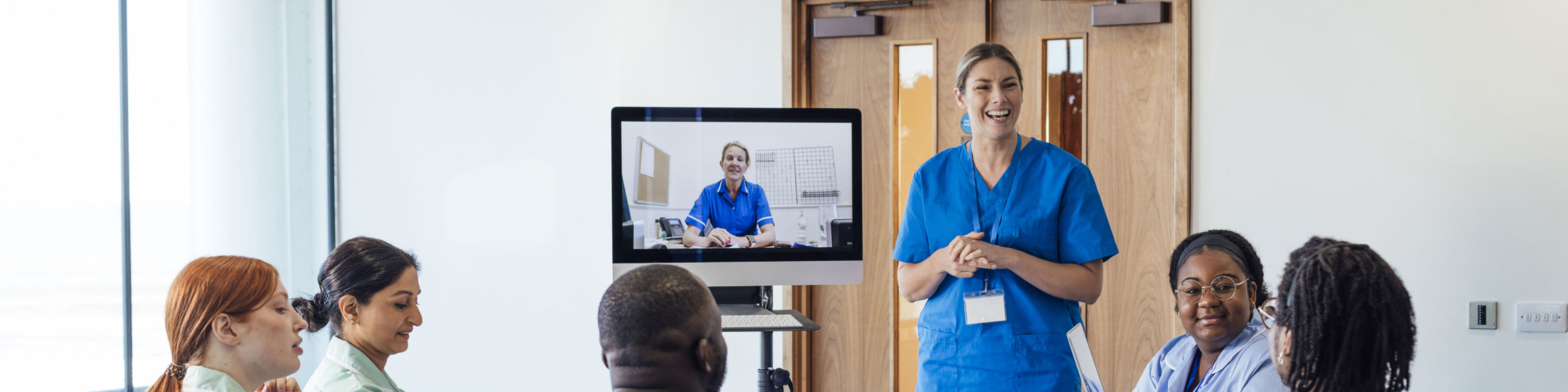 A group of healthcare professionals sit around a table, listening to a colleague in blue scrubs who is standing and presenting. Another person in scrubs appears on a monitor, participating via video call.