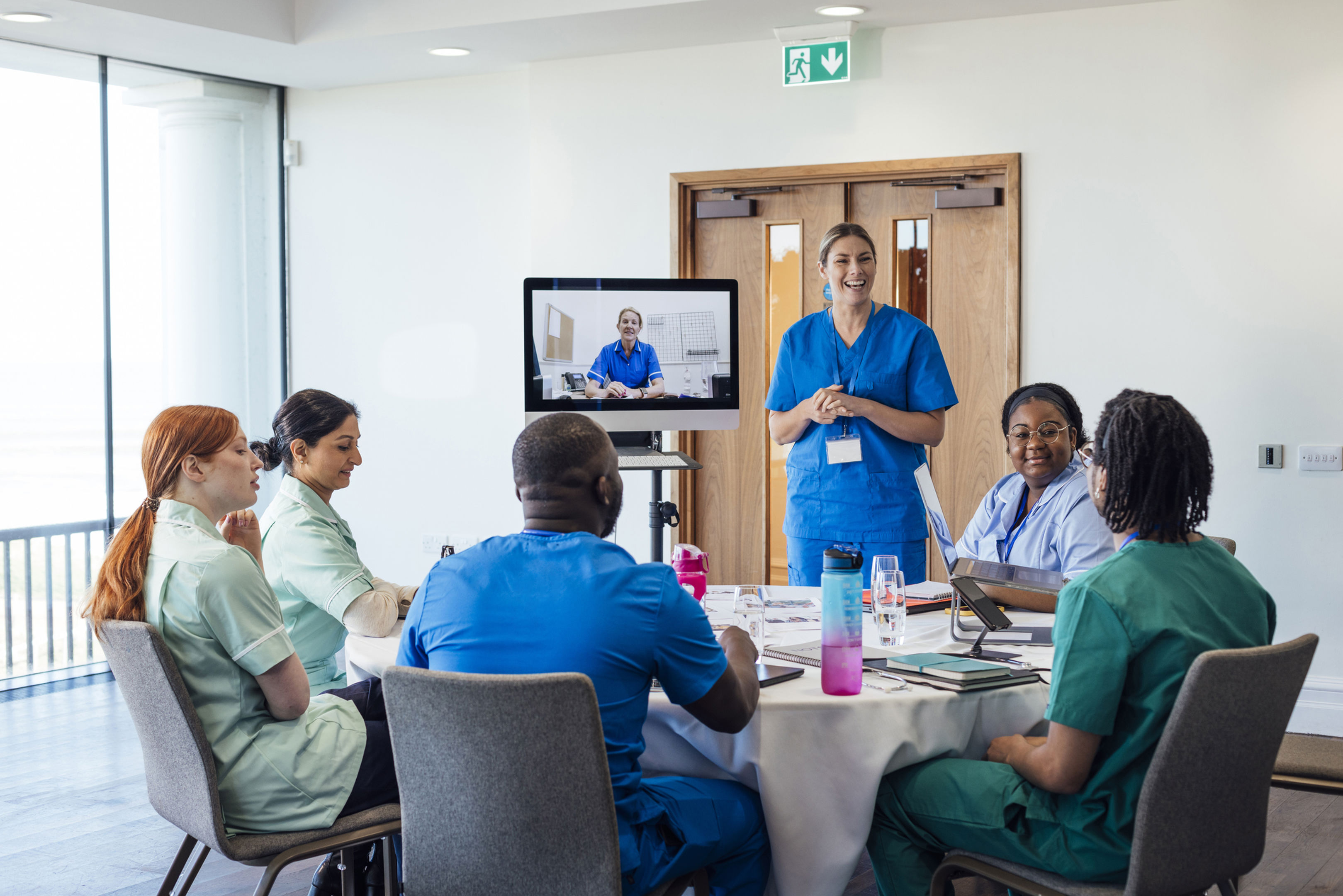 A group of healthcare professionals sit around a table, listening to a colleague in blue scrubs who is standing and presenting. Another person in scrubs appears on a monitor, participating via video call.