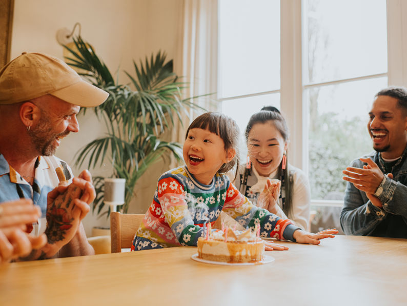 A smiling child sits at a table with a birthday cake while three adults around them clap and laugh, celebrating together in a bright room.