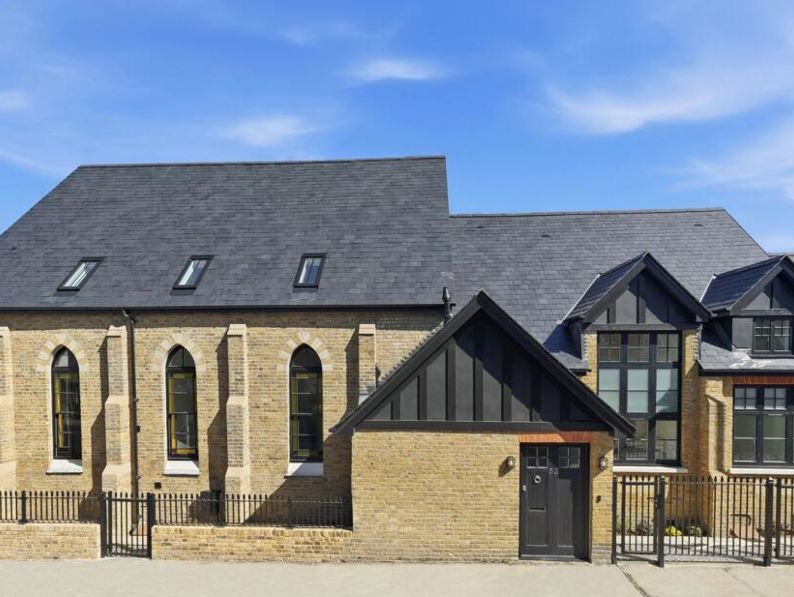 A modernized building with a steep slate roof, brick walls, arched windows, and black trim, resembling a converted church or historical structure under a blue sky.