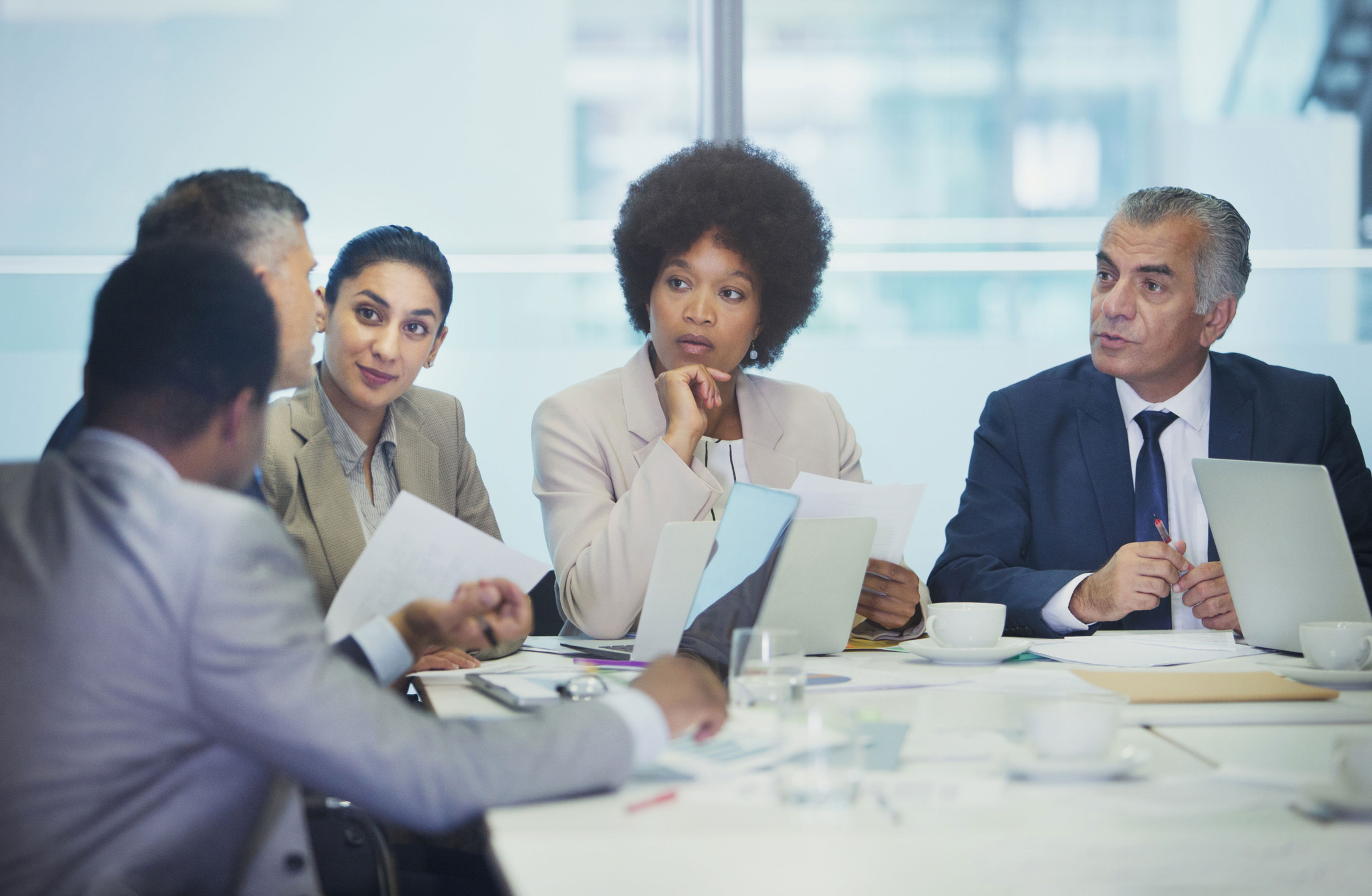 Five professionals sit around a conference table in discussion, holding documents and laptops. They appear focused and engaged in a meeting, with coffee cups and office materials on the table.