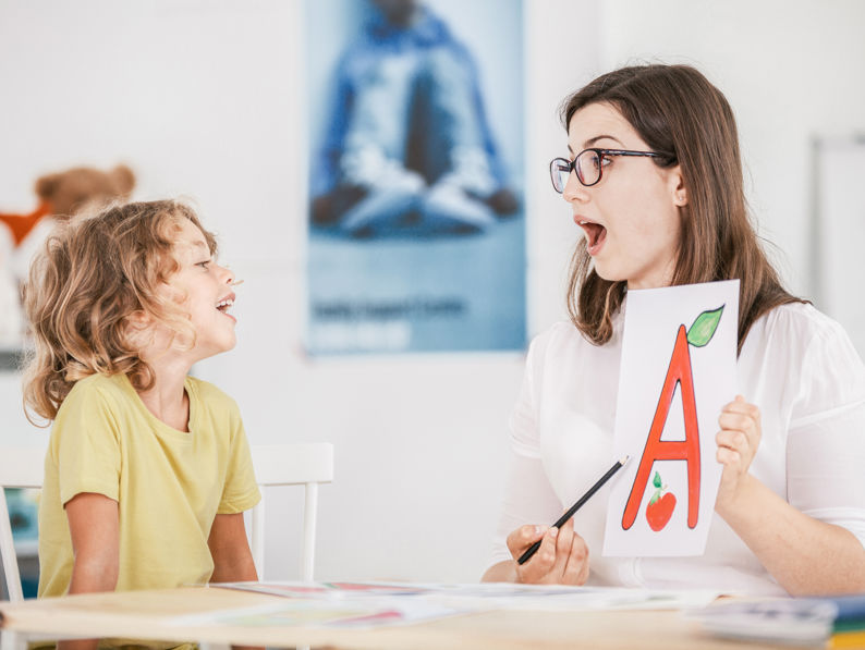 A teacher holding up a paper with a large letter “A” and pictures of an apple and leaf, sitting at a table with a smiling child in a classroom setting.