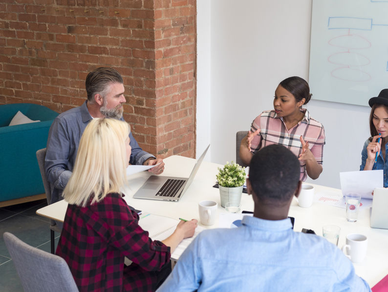 Five people sit around a table in a modern office, engaged in discussion with laptops, papers, and coffee cups in front of them. A whiteboard with diagrams is visible in the background.