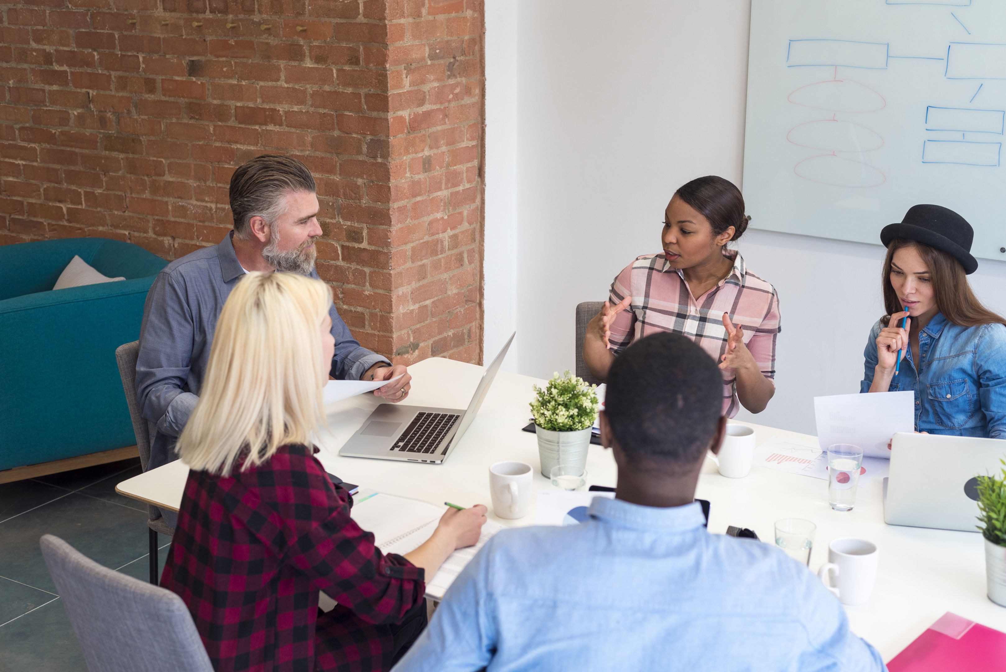 Five people sit around a table in a modern office, engaged in discussion with laptops, papers, and coffee cups in front of them. A whiteboard with diagrams is visible in the background.