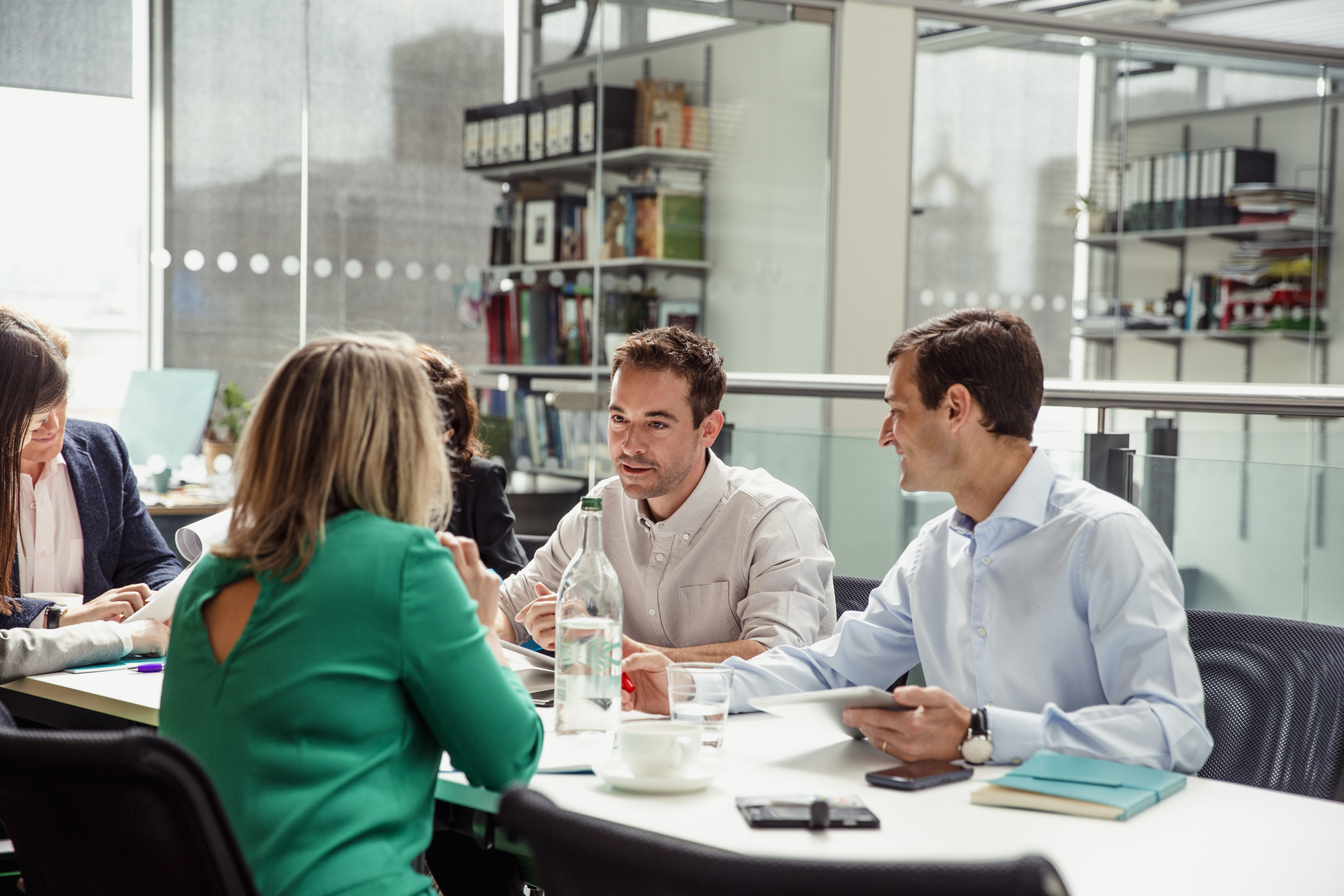 Four people sit around a table in a modern office, engaged in discussion. Papers, notebooks, and a water bottle are on the table, and shelves with books and files are visible in the background.